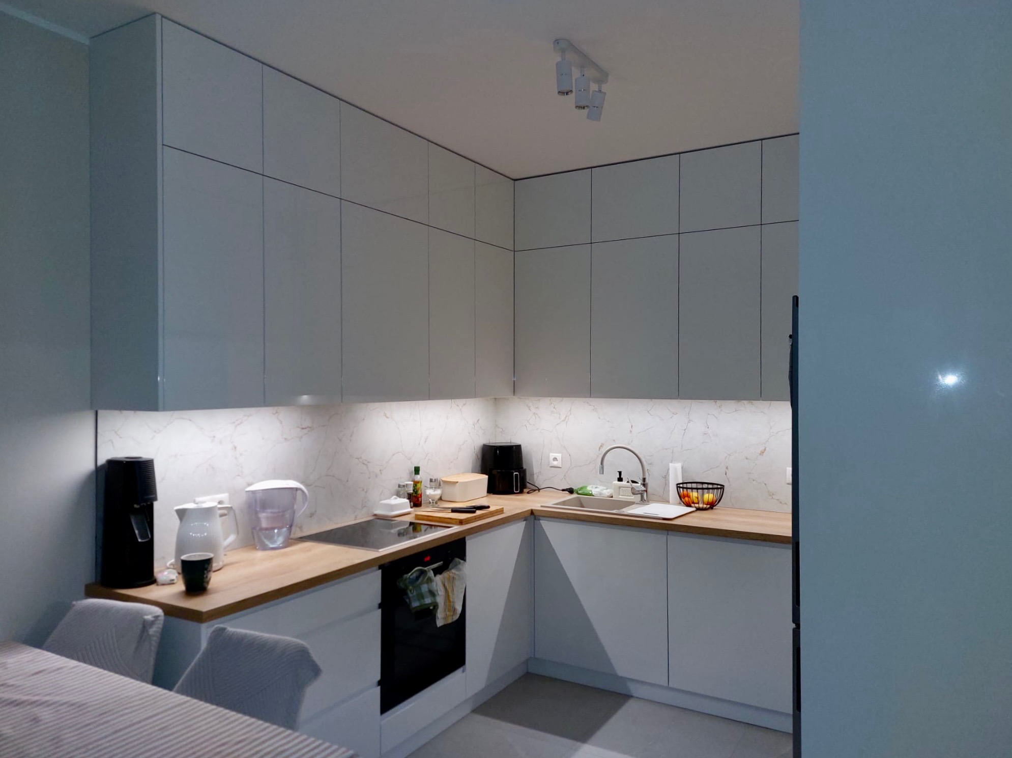Jasna kuchnia z białymi, gładkimi frontami szafek i drewnianym blatem. Uwagę zwraca marmurowy wzór na ścianie nad blatem i nowoczesne oświetlenie podszafkowe.