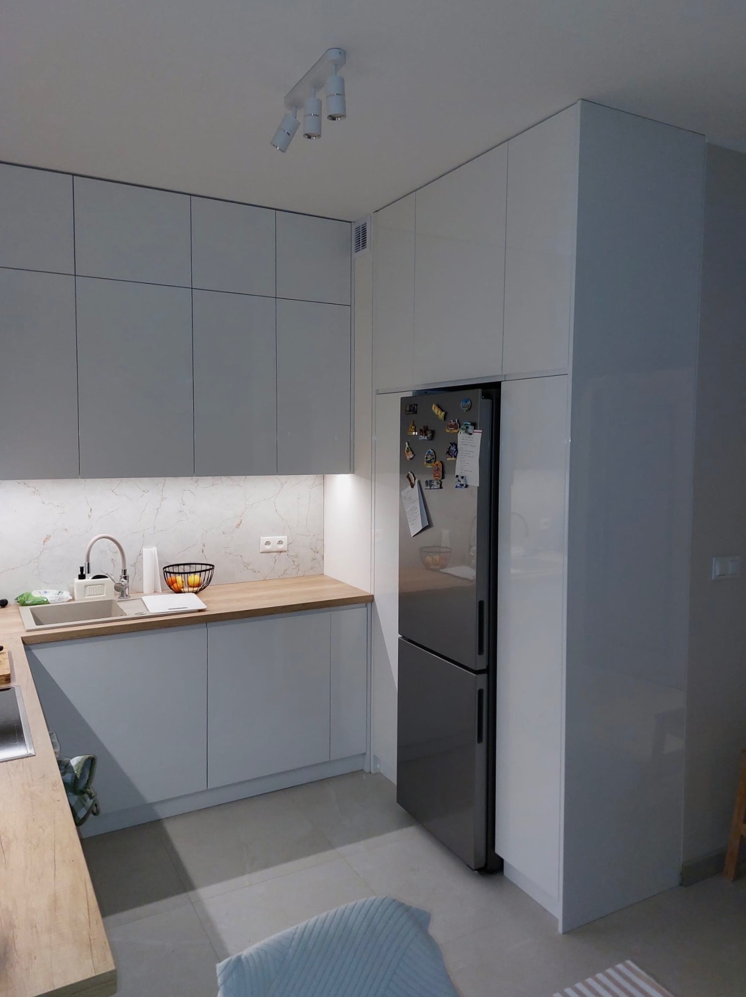 Nowoczesna kuchnia z szarymi, gładkimi frontami szafek, drewnianym blatem i marmurowym backsplashiem. Lodówka w zabudowie z magnesami. Oświetlenie punktowe.