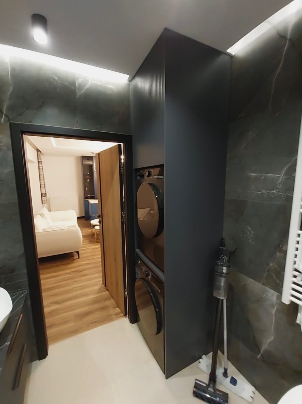 Nowoczesna zabudowa łazienkowa z pralką i suszarką w kolumnie, ciemnoszara szafa z ryflowanym frontem, marmurowe ściany, widok na salon przez otwarte drzwi.