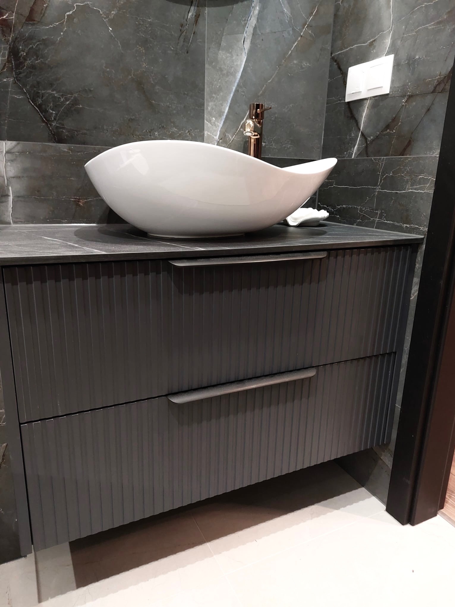 Szara szafka łazienkowa z umywalką w kształcie łodzi na blacie z ciemnego kamienia, na tle ściany z marmuru. Widoczne chromowane detale i minimalistyczny design.