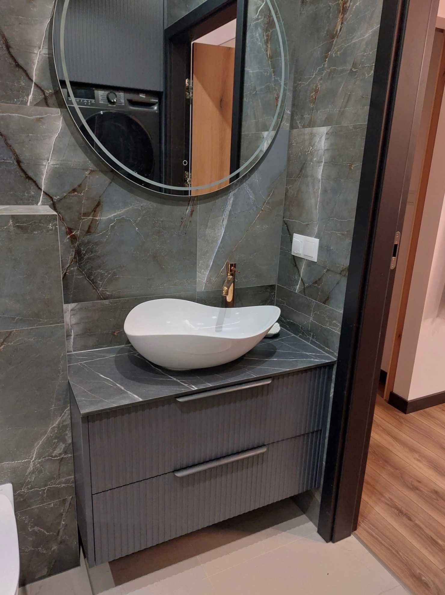 Nowoczesna łazienka z umywalką nablatową w kształcie łzy, szafką z frezowanymi frontami i lustrem. Marmurowe płytki na ścianach i złota bateria.