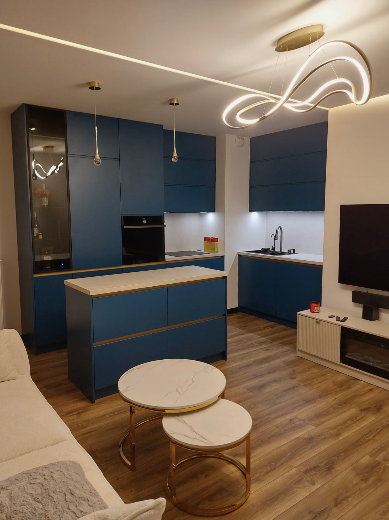 Niebieska kuchnia z wyspą i złotymi akcentami, nowoczesne oświetlenie, widok na salon z sofą i stolikami kawowymi. Całość w ciepłej tonacji.