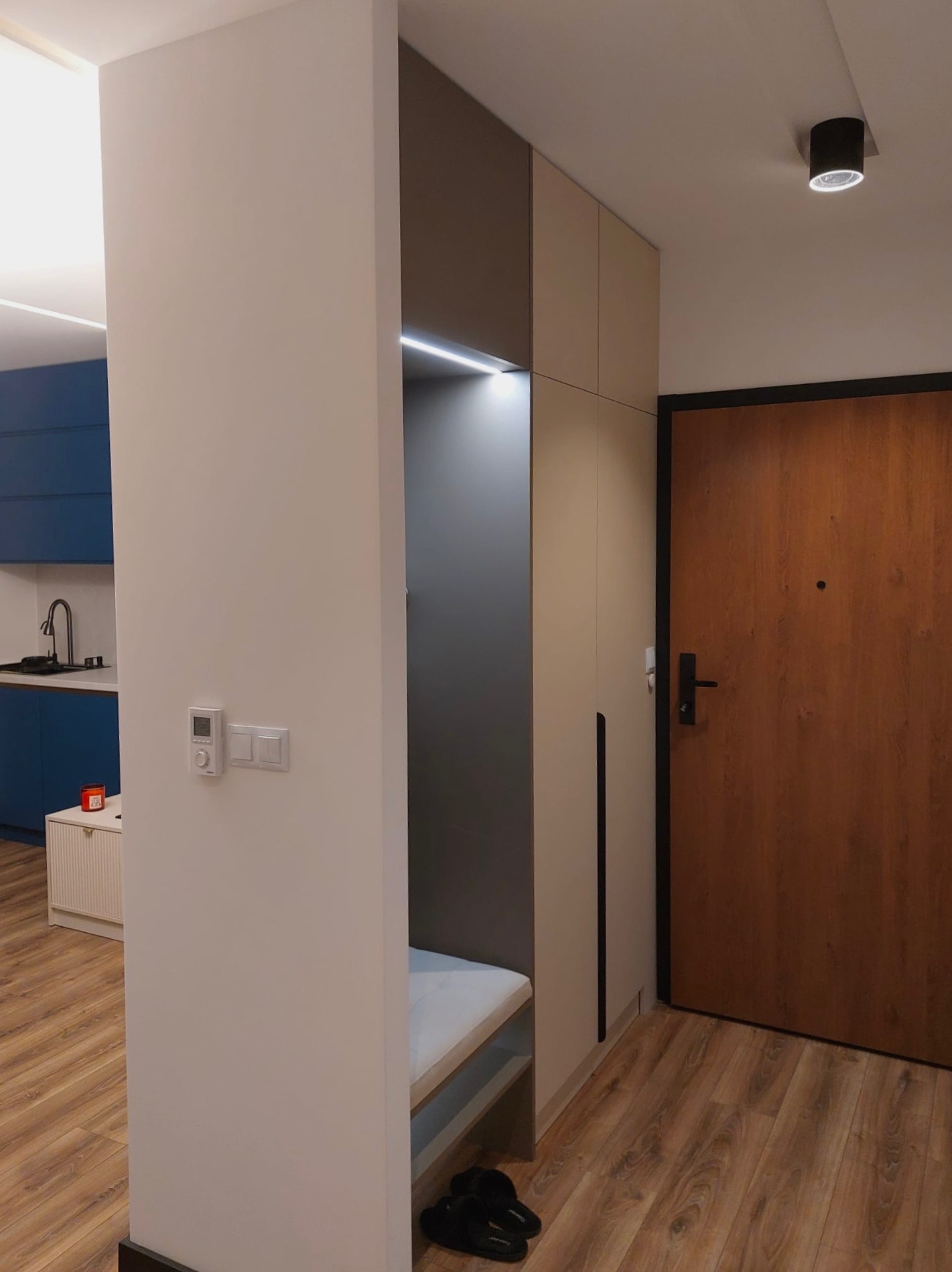 Nowoczesna szafa wnękowa z siedziskiem w przedpokoju. Minimalistyczny design, oświetlenie LED, drewniana podłoga i drzwi. Widoczny fragment niebieskiej kuchni.