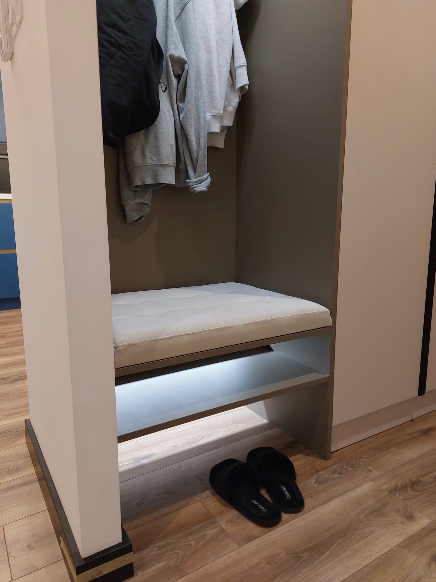 Szafa wnękowa z siedziskiem i oświetleniem LED, w kolorach szarości i bieli. Na półce pod siedziskiem leżą czarne kapcie. U góry wieszaki z ubraniami.