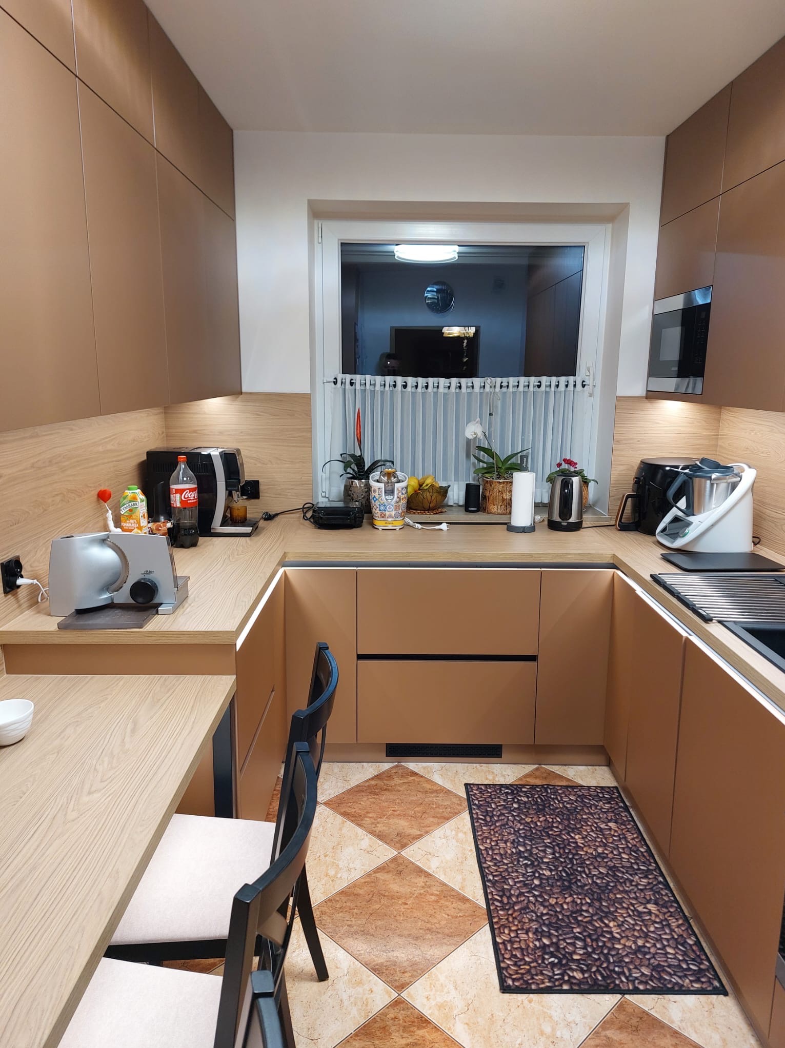 Nowoczesna kuchnia w kształcie litery U z beżowymi frontami, drewnianym blatem i oświetleniem LED. Na blacie ekspres do kawy i inne akcesoria. Na podłodze dywanik z motywem kawy.