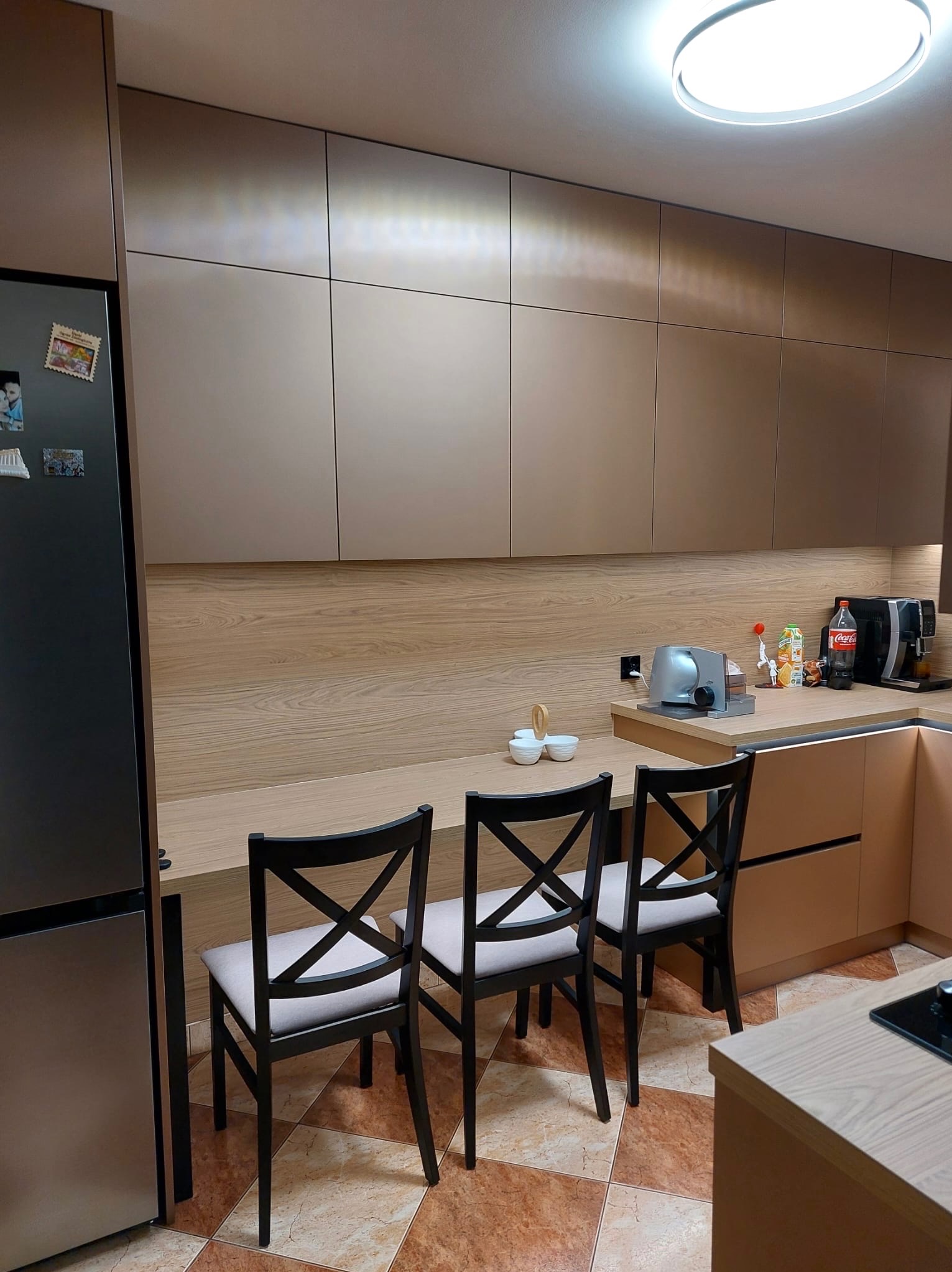 Nowoczesna kuchnia na wymiar z zabudową w odcieniach beżu i drewna, zintegrowana z blatem pełniącym funkcję barku i trzema krzesłami. Widoczny sprzęt AGD i akcesoria.