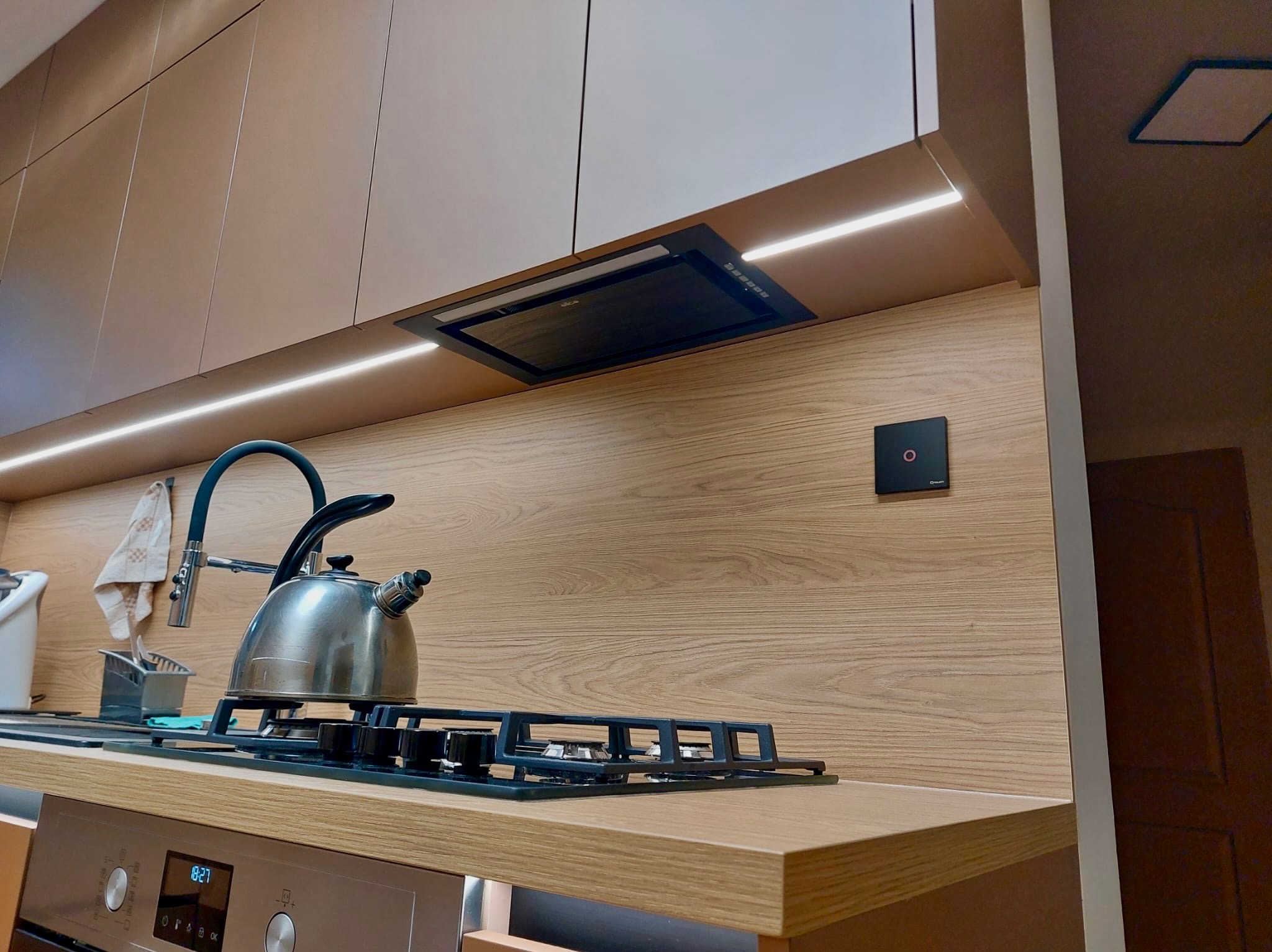 Nowoczesna kuchnia z drewnianym blatem i oświetleniem LED w Radomsku. Na blacie czajnik i płyta gazowa, nad nimi okap. Jasne fronty szafek górnych.