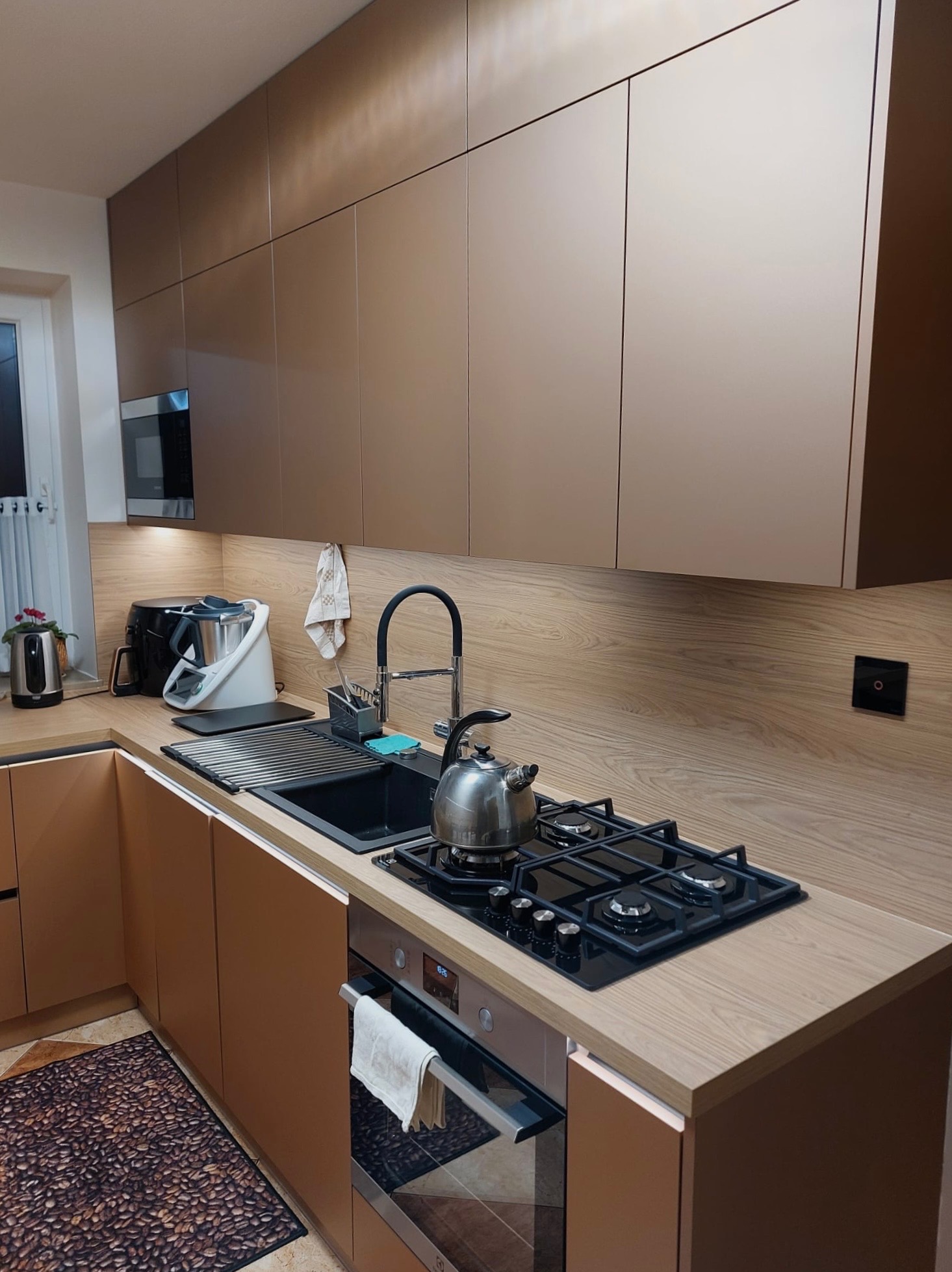 Nowoczesna kuchnia z zabudową w odcieniach beżu i drewna. Górne szafki bez uchwytów, czarny zlew i płyta gazowa. Na blacie czajnik i akcesoria kuchenne.
