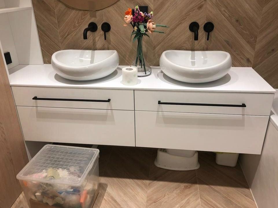 Biała szafka łazienkowa z dwiema umywalkami nablatowymi w kształcie misy i czarną armaturą, uzupełniona o wazon z kwiatami i akcesoria łazienkowe. Pod szafką widoczny pojemnik z akcesoriami...