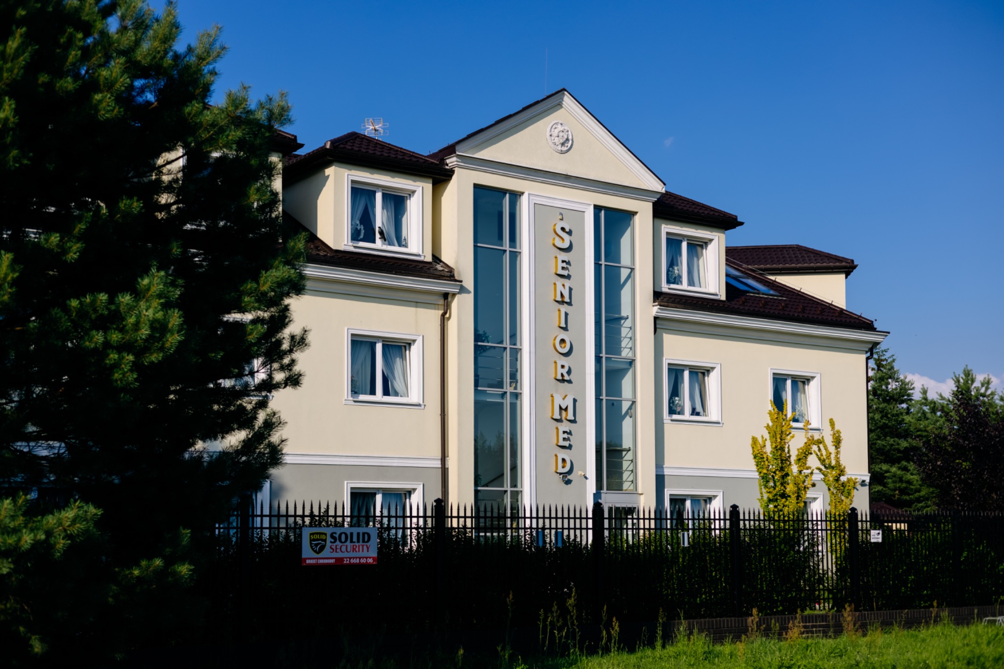 Budynek domu opieki Senior Med z napisem na fasadzie, widoczny zza ogrodzenia i zieleni. Na pierwszym planie tabliczka Solid Security.