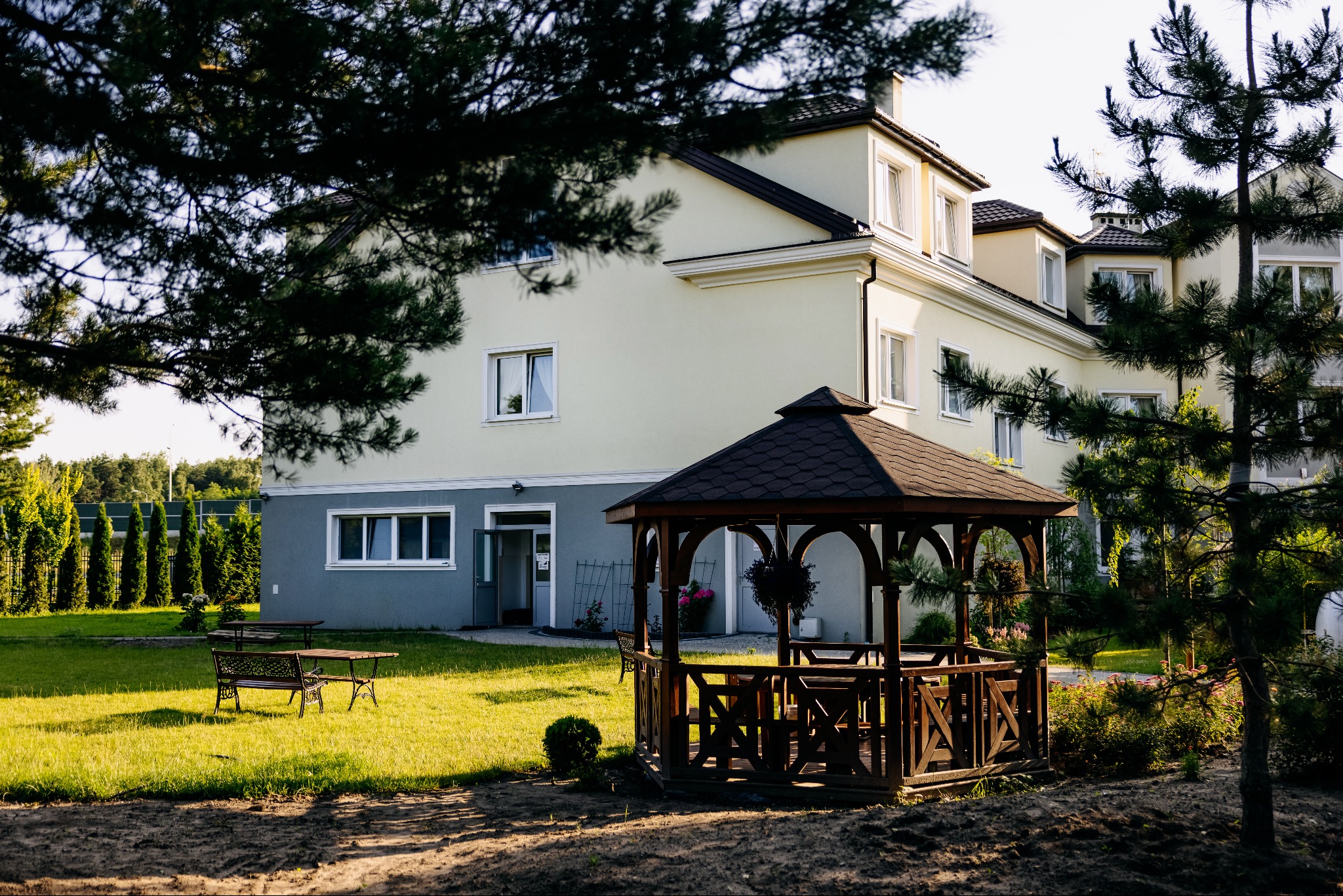Dwupiętrowy budynek domu opieki z ogrodem, altaną i ławkami. Jasna elewacja, ciemny dach. Widok z zewnątrz, drzewa w kadrze. Słoneczny dzień.