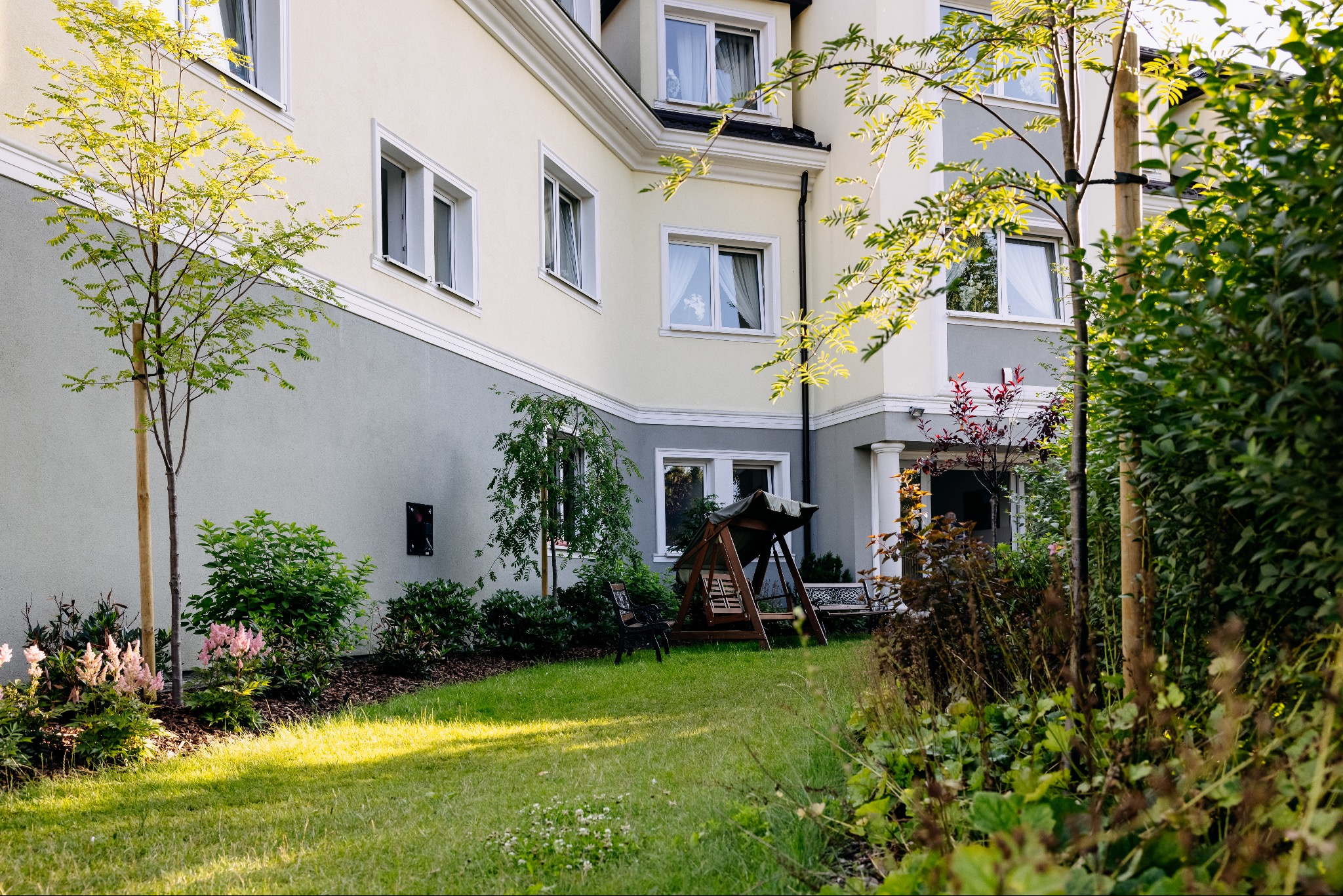 Ogród z trawnikiem i roślinnością przed budynkiem domu opieki, z widocznym wejściem i huśtawką ogrodową. Jasna elewacja budynku z szarymi akcentami.