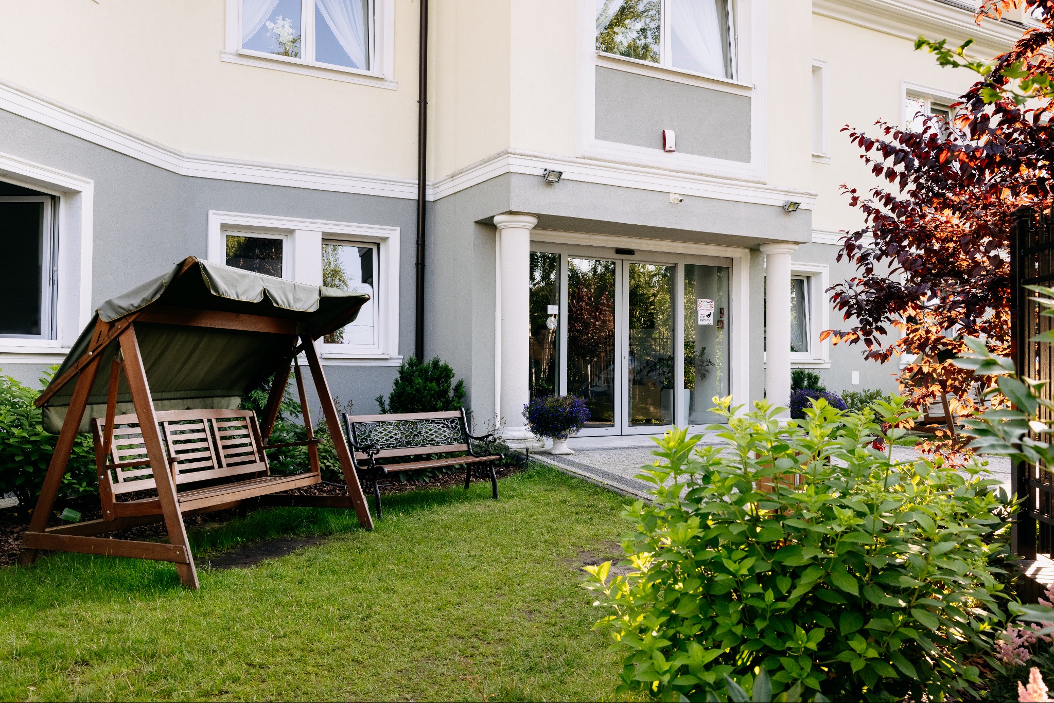 Elewacja budynku z ogrodem, huśtawką i ławką. Wejście z kolumnami i automatycznymi drzwiami. Zieleń, krzewy i drzewa. Dom opieki z miejscem do wypoczynku na zewnątrz.
