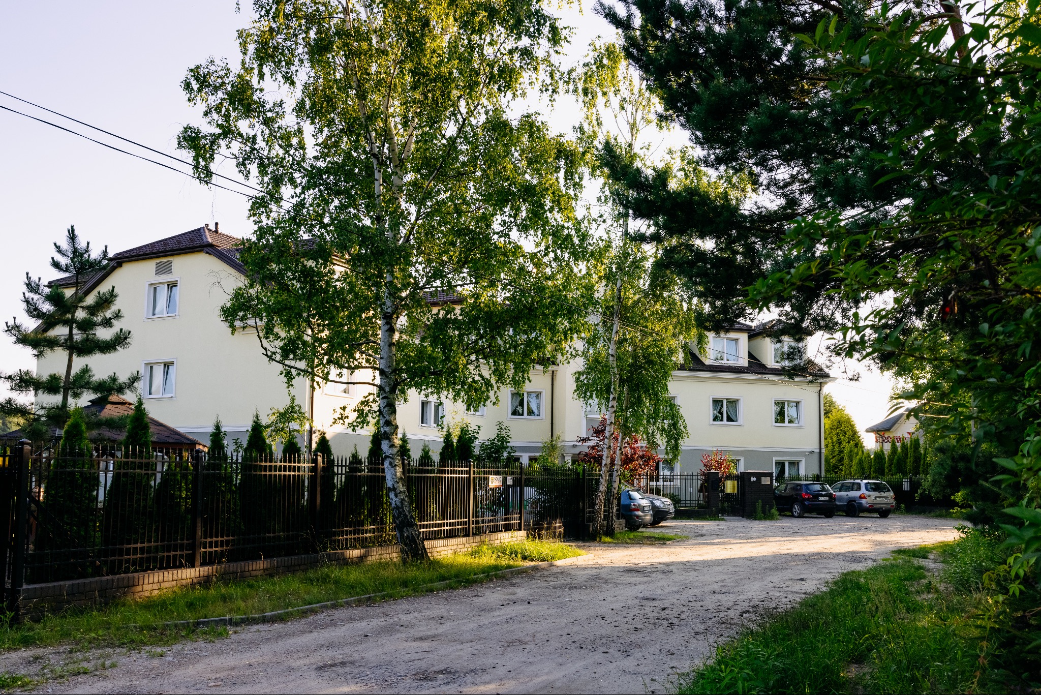Jasny budynek domu opieki z podjazdem i zaparkowanymi autami, widok z ulicy przez metalowe ogrodzenie i drzewa. Ujęcie w słoneczny dzień.