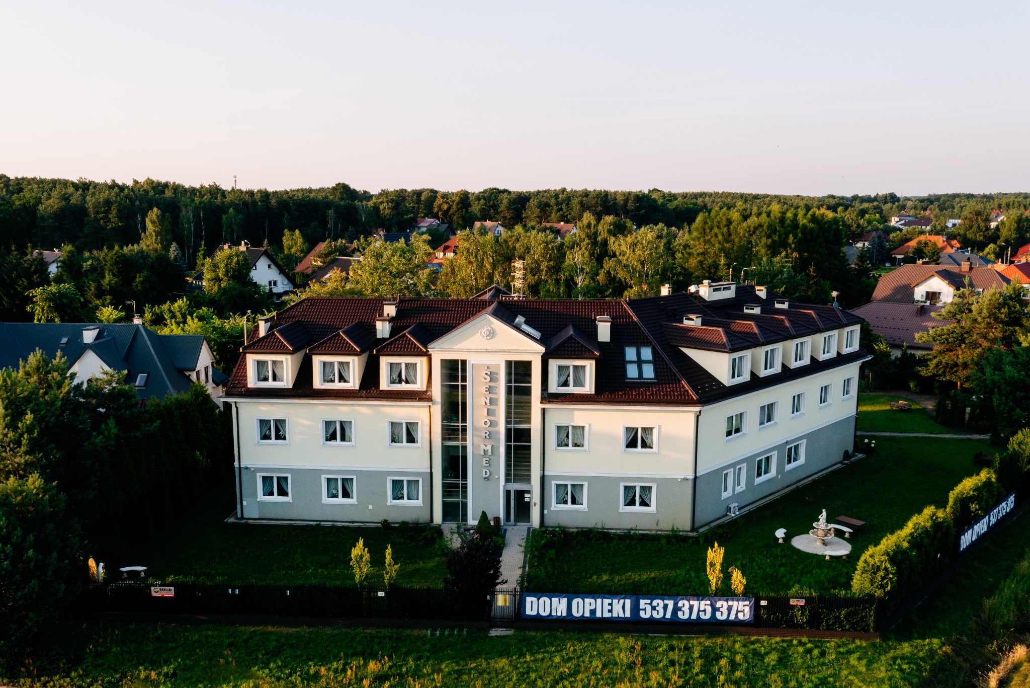 Dom opieki Senior Med z ogrodem, widok z góry. Budynek z brązowym dachem, jasną elewacją i dużymi oknami. Na ogrodzeniu baner z numerem telefonu.