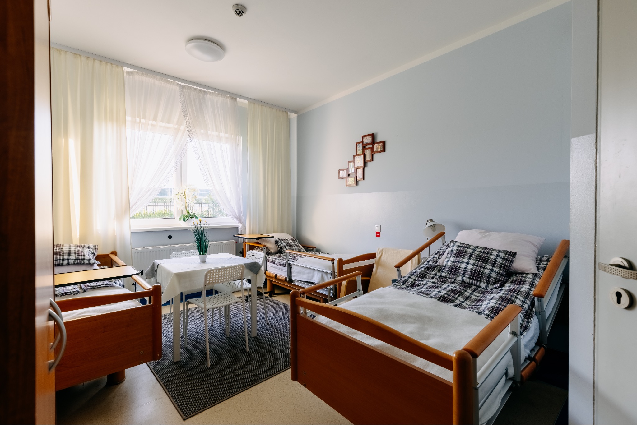 Jasny, przestronny pokój w domu opieki z trzema łóżkami, stolikiem i kwiatami. Widok na okno z firankami. Ustawione zdjęcia na ścianie. Przytulne wnętrze.