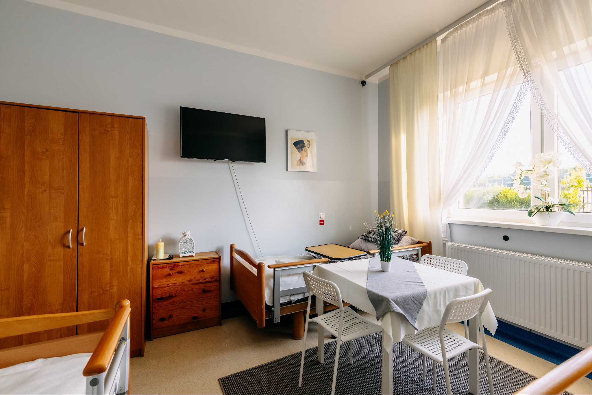 Pokój w domu seniora w Wiązownie: łóżko rehabilitacyjne, stół z krzesłami, szafa, komoda, telewizor na ścianie, okno z widokiem na zieleń. Jasne, przytulne wnętrze.