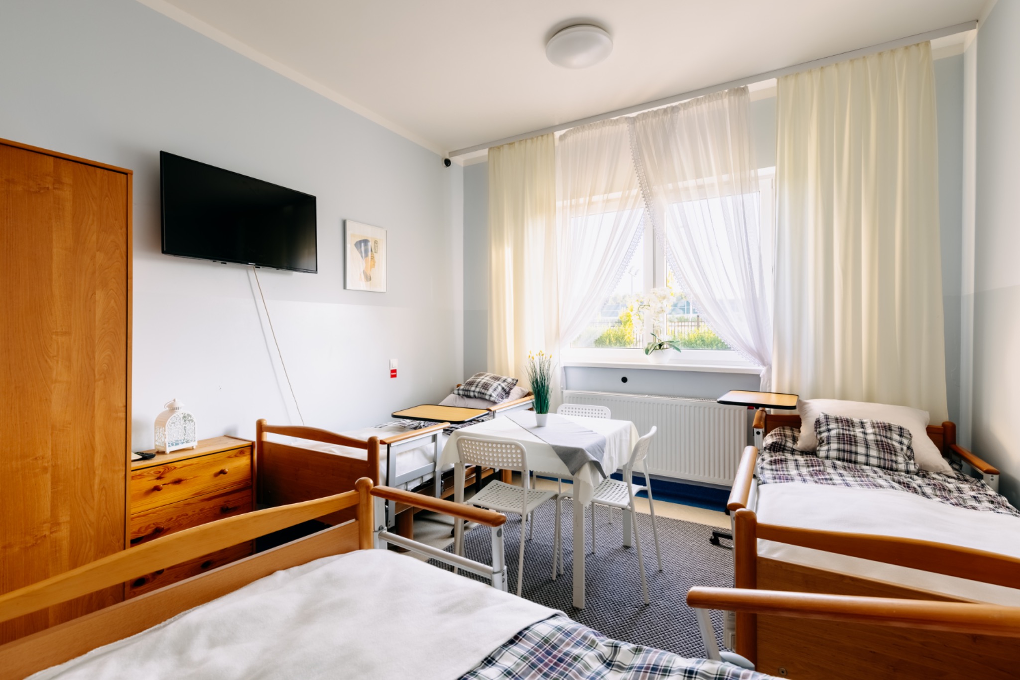Jasny pokój w domu opieki: dwa łóżka z drewnianymi ramami, stolik z krzesłami, telewizor na ścianie, okno z firankami. Przytulne wnętrze dla seniorów.