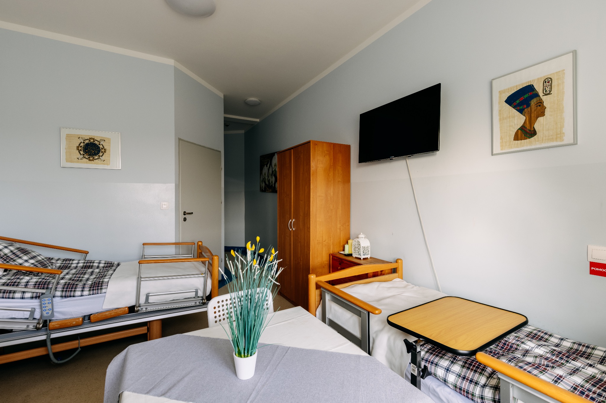 Pokój w domu opieki: dwa łóżka z regulacją, szafa, stolik, telewizor i dekoracje ścienne. Jasne, przestronne wnętrze z elementami wyposażenia dla seniorów.