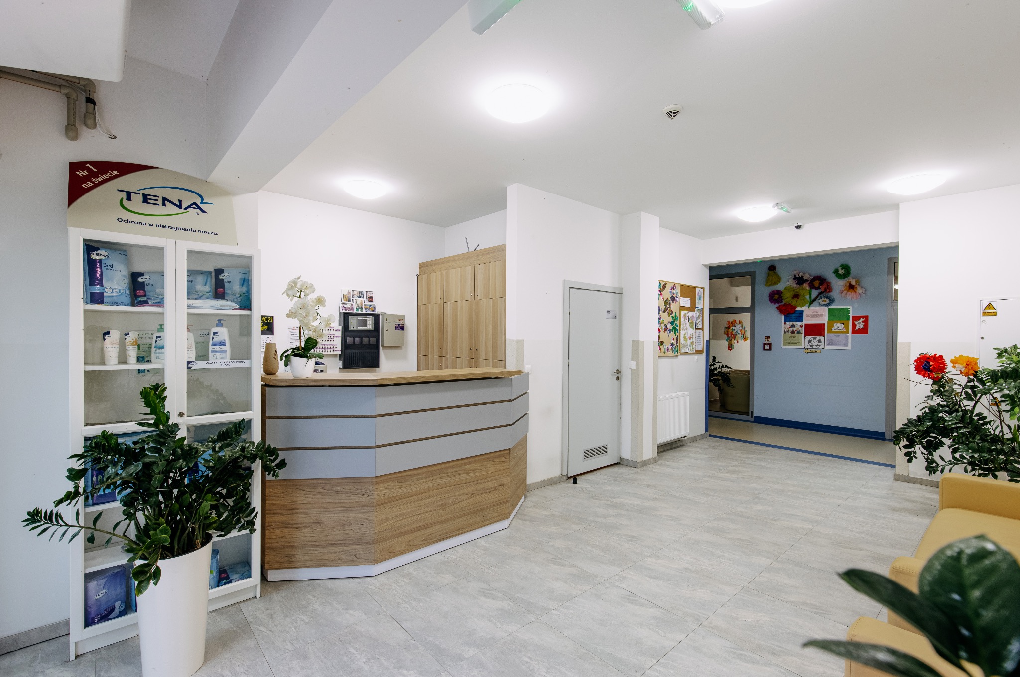 Wnętrze domu opieki: recepcja z drewnianym wykończeniem, regał z produktami higienicznymi TENA, kwiaty i dekoracje na ścianach. Jasne, przestronne pomieszczenie.