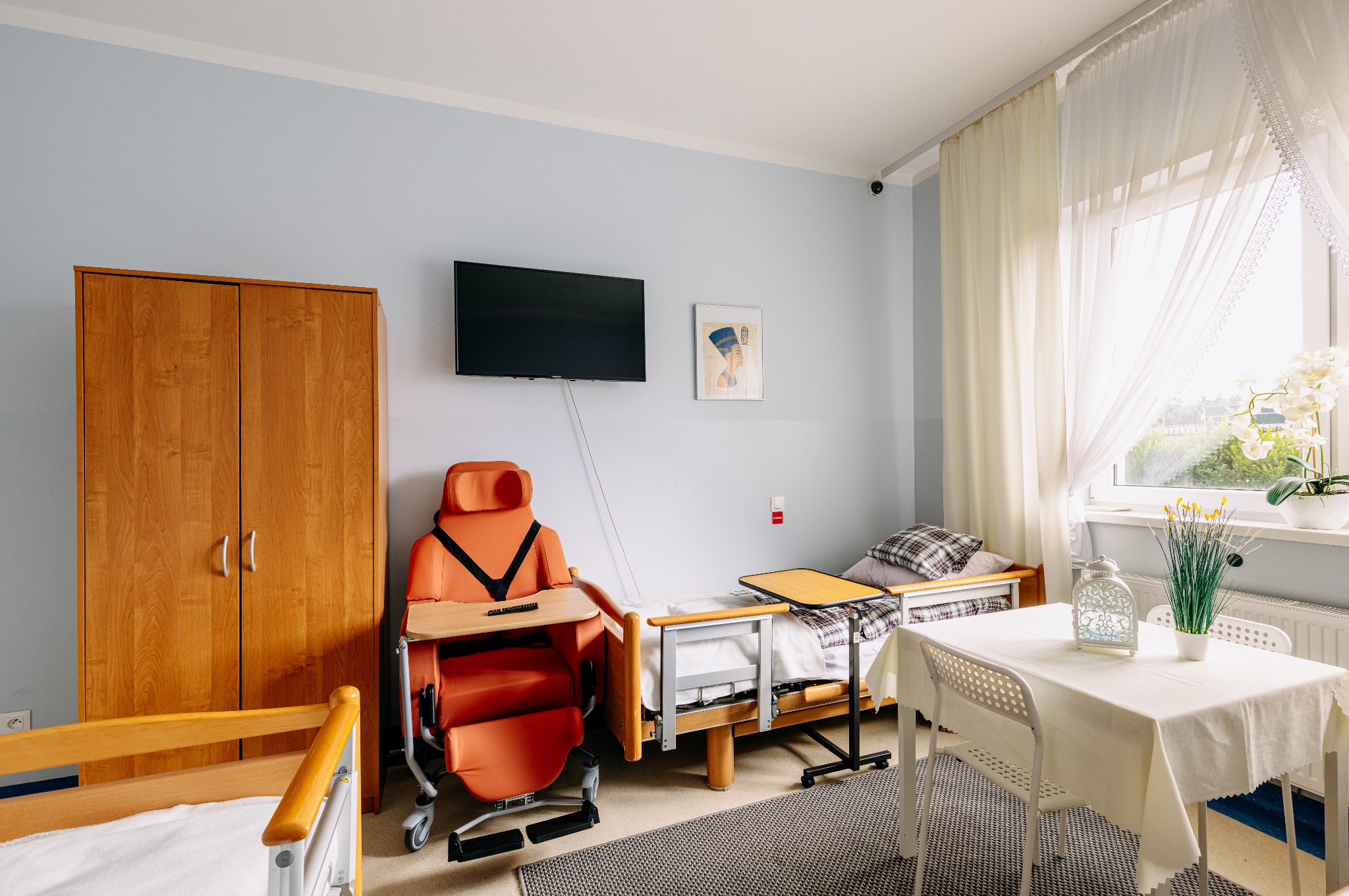 Pokój w domu opieki: łóżko z szafką, fotel rehabilitacyjny, stół z dekoracją okna. Jasne wnętrze, spokojna kolorystyka, zapewniające komfortowe warunki pobytu.