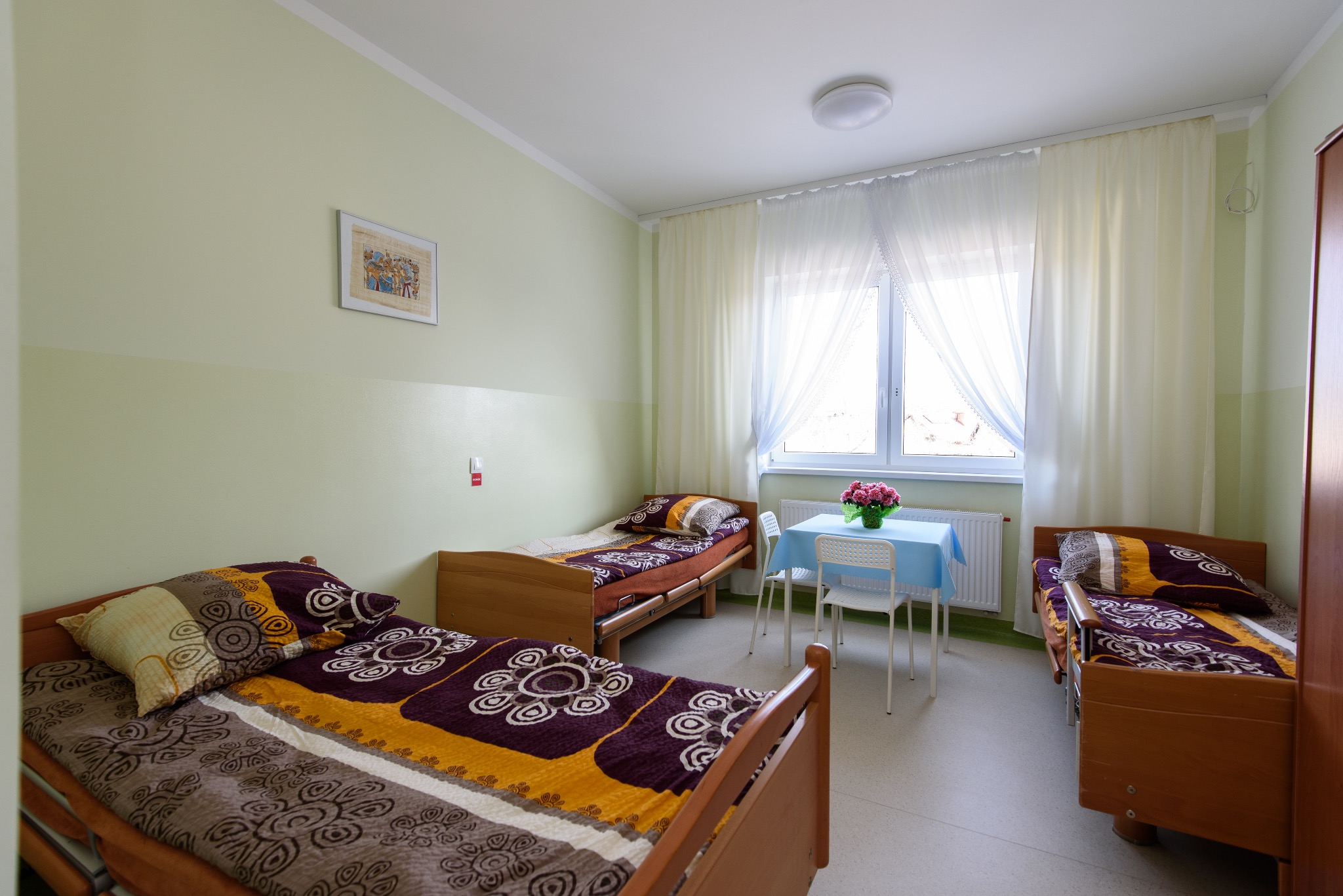 Jasny pokój w placówce opiekuńczej z trzema łóżkami z wzorzystą pościelą, stolikiem z kwiatami pod oknem z białymi firankami i zielonymi ścianami.