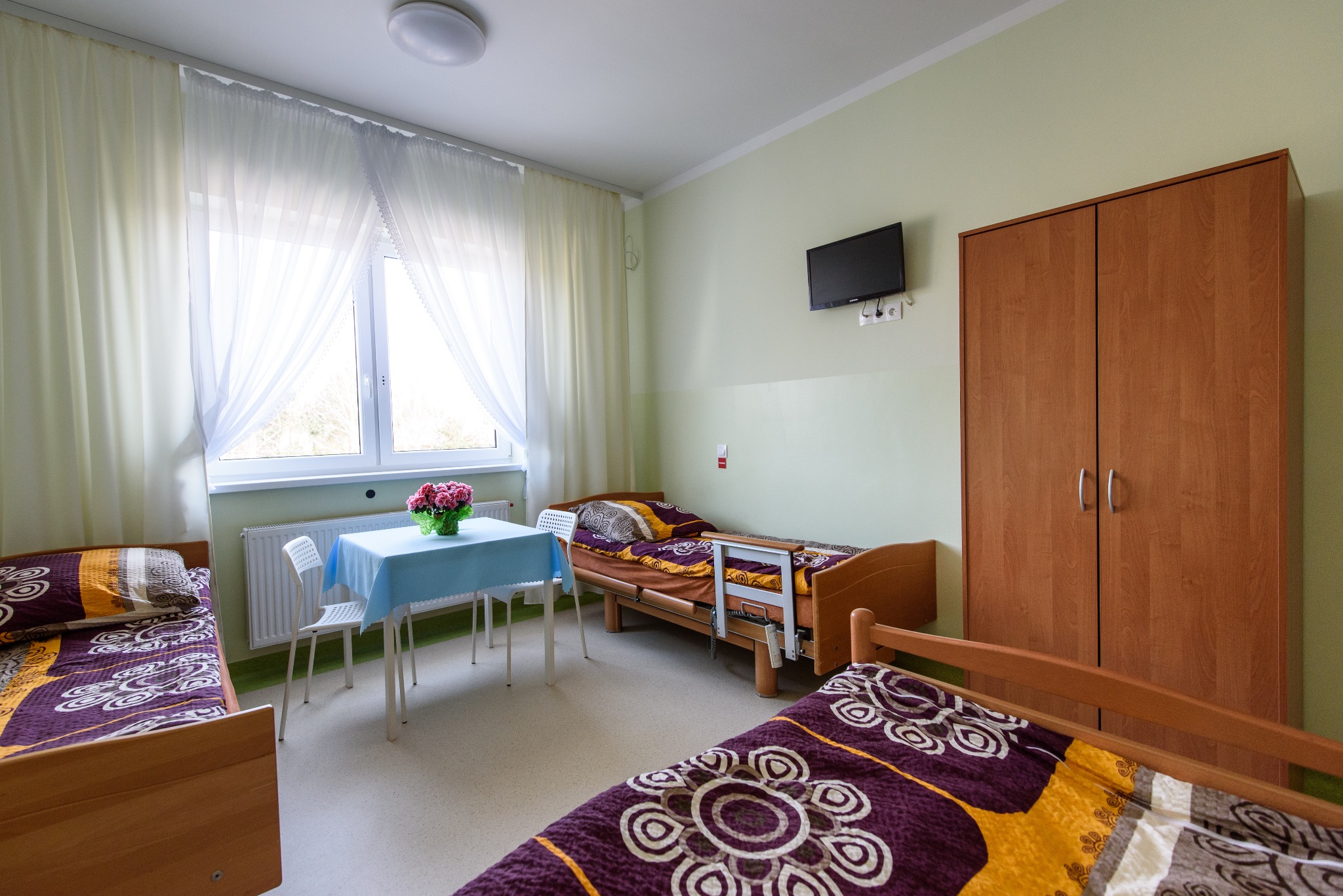 Jasny pokój w domu opieki: trzy łóżka z kolorową pościelą, stół z kwiatami, telewizor na ścianie i drewniana szafa. Okno z białymi firankami.