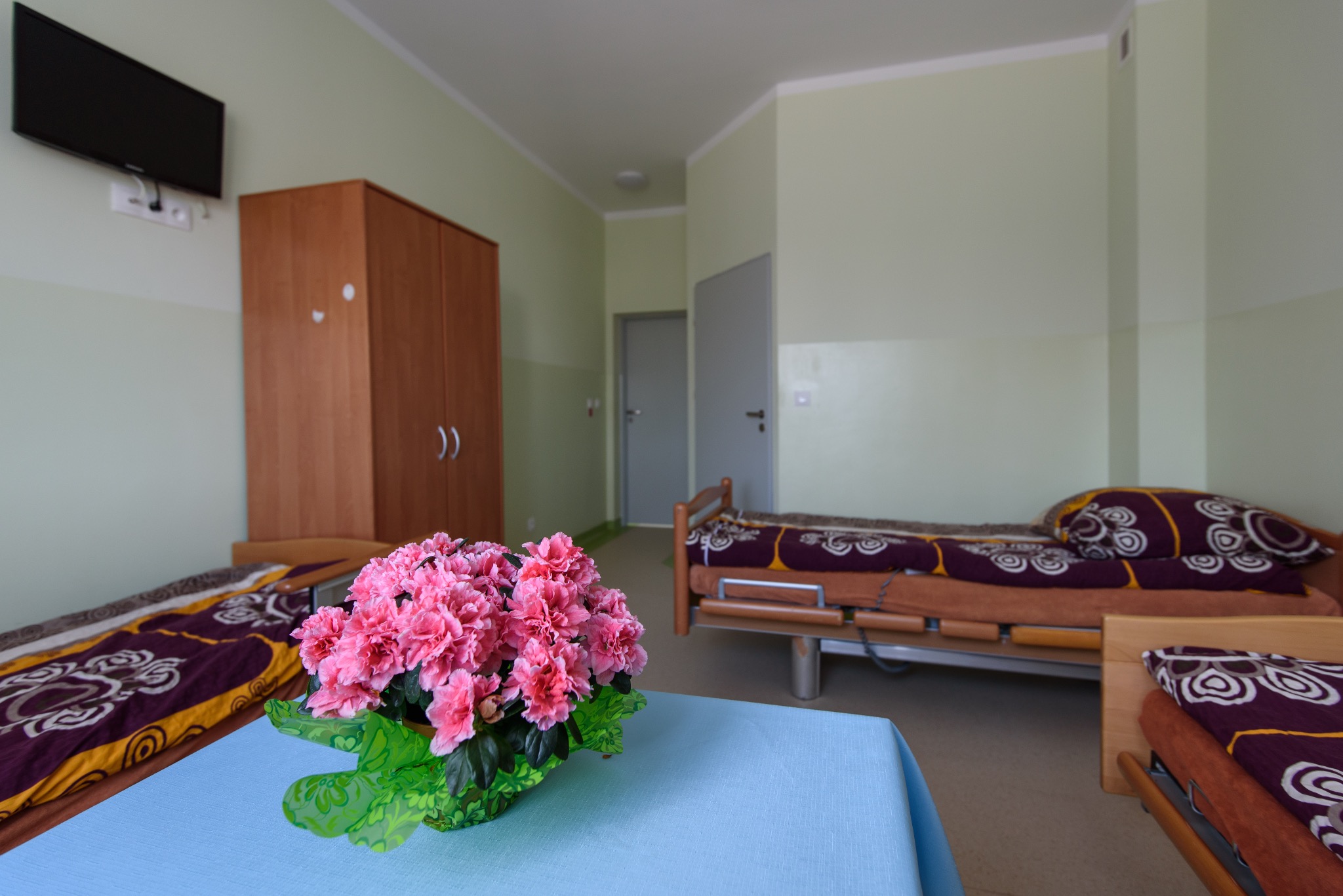Pokój w domu opieki z trzema łóżkami, szafą i telewizorem. Na pierwszym planie stół z bukietem różowych kwiatów. Jasne ściany i proste wyposażenie.