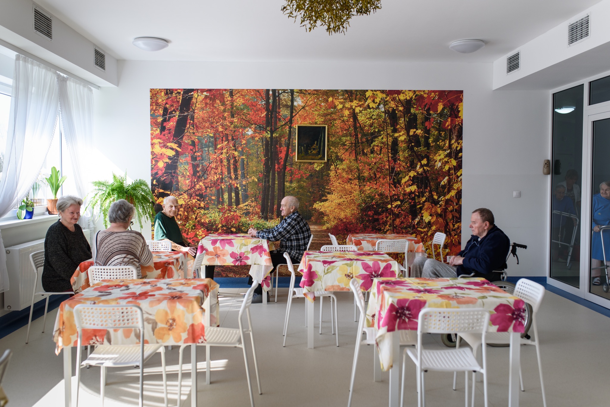 Jasna jadalnia w domu opieki z mieszkańcami siedzącymi przy stolikach z kwiatowymi obrusami, na ścianie fototapeta z jesiennym lasem.