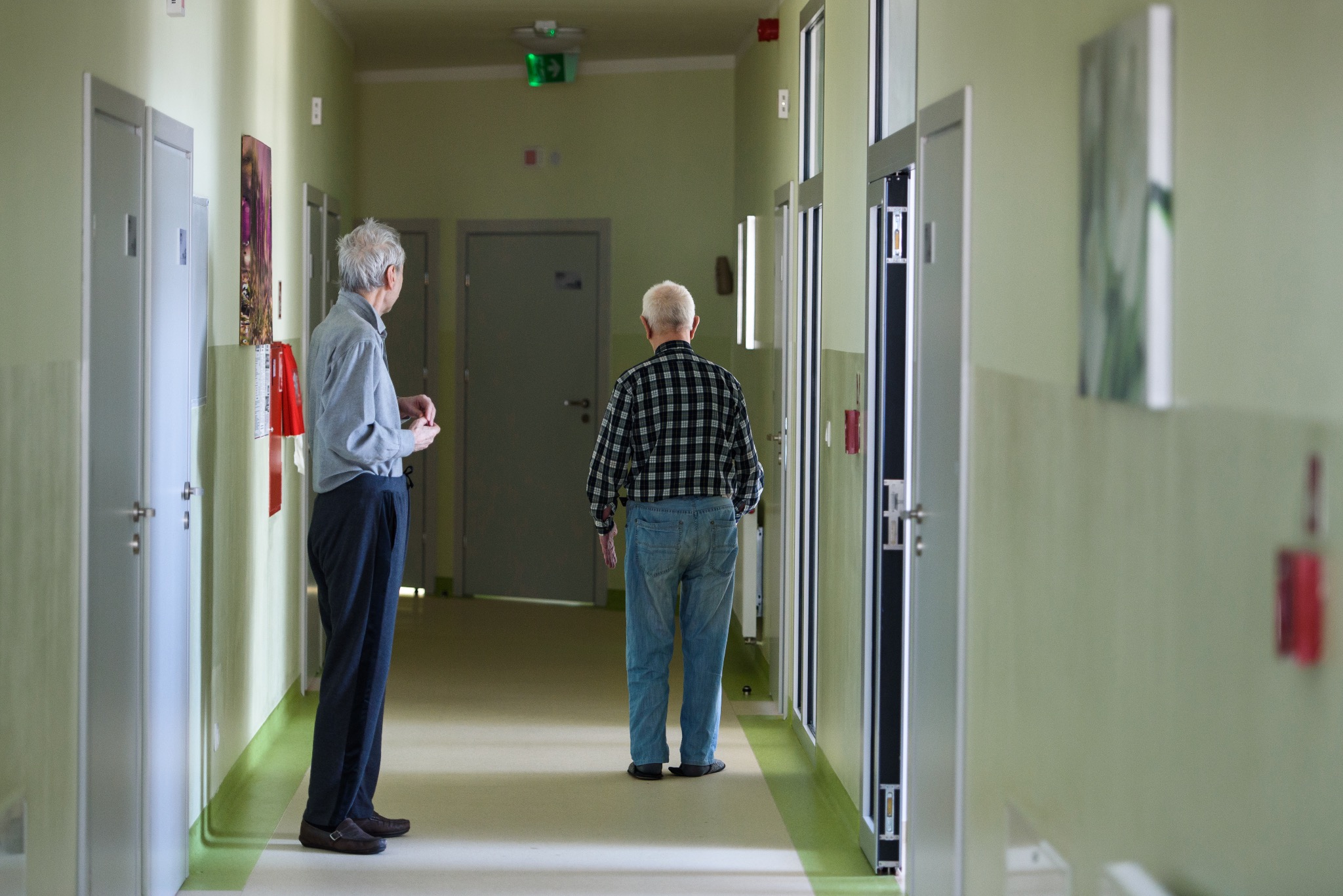 Korytarz w domu opieki: Dwóch seniorów stoi w jasnym korytarzu z zielonymi ścianami i białymi drzwiami. Jeden patrzy w dal, drugi stoi tyłem. Oświetlenie sufitowe.