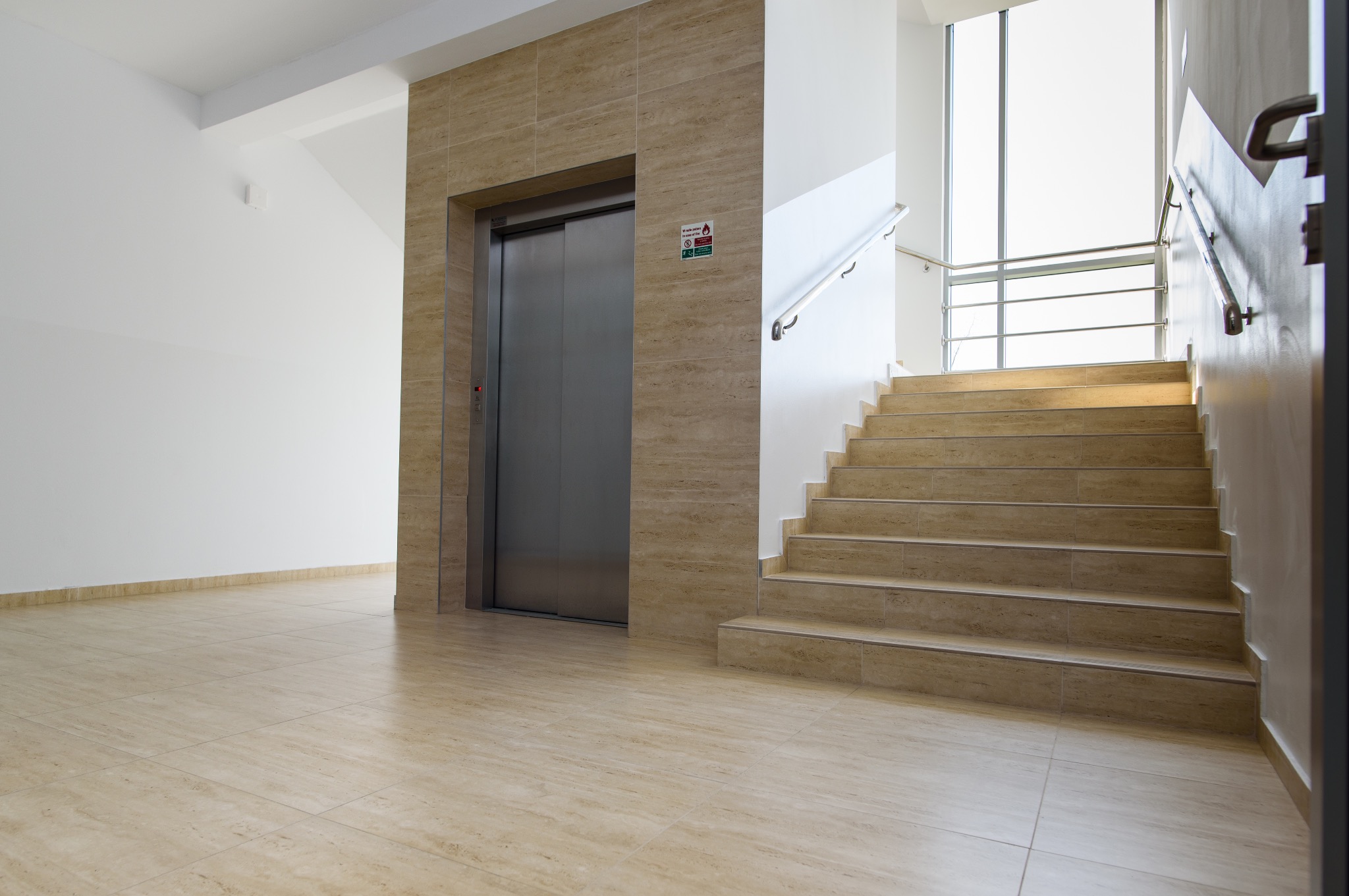 Jasne wnętrze z beżowymi płytkami podłogowymi, schodami z poręczą i windą. Widoczny znak informacyjny na ścianie obok windy. Dostępność dla osób z ograniczeniami ruchowymi.