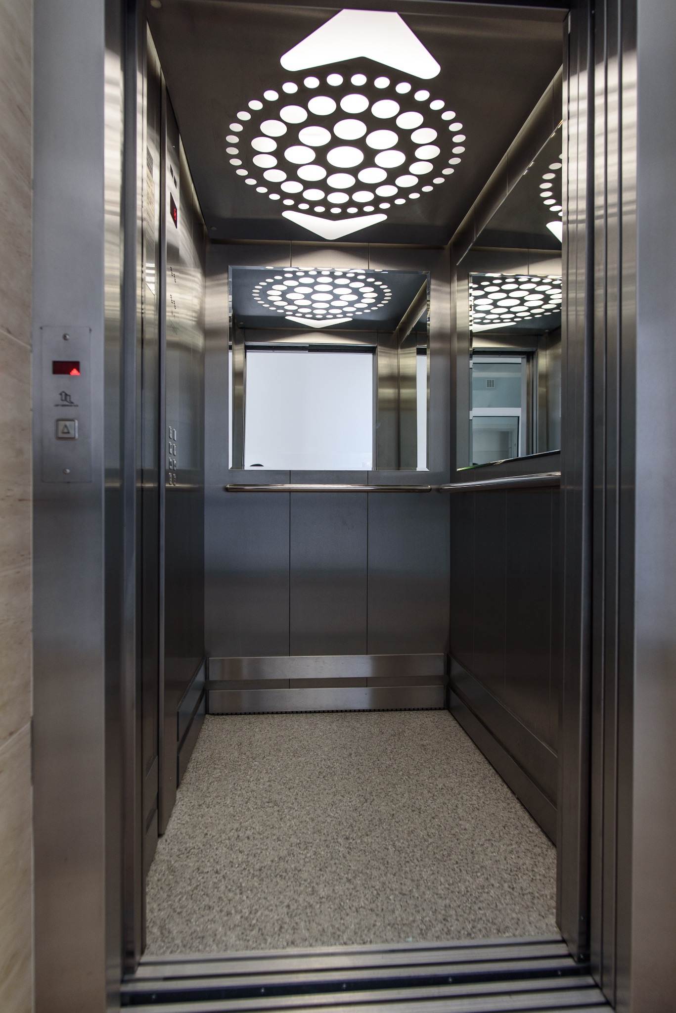 Wnętrze nowoczesnej windy w domu opieki, z lustrem, oświetleniem sufitowym w kształcie rozety i panelami sterowania po lewej stronie.