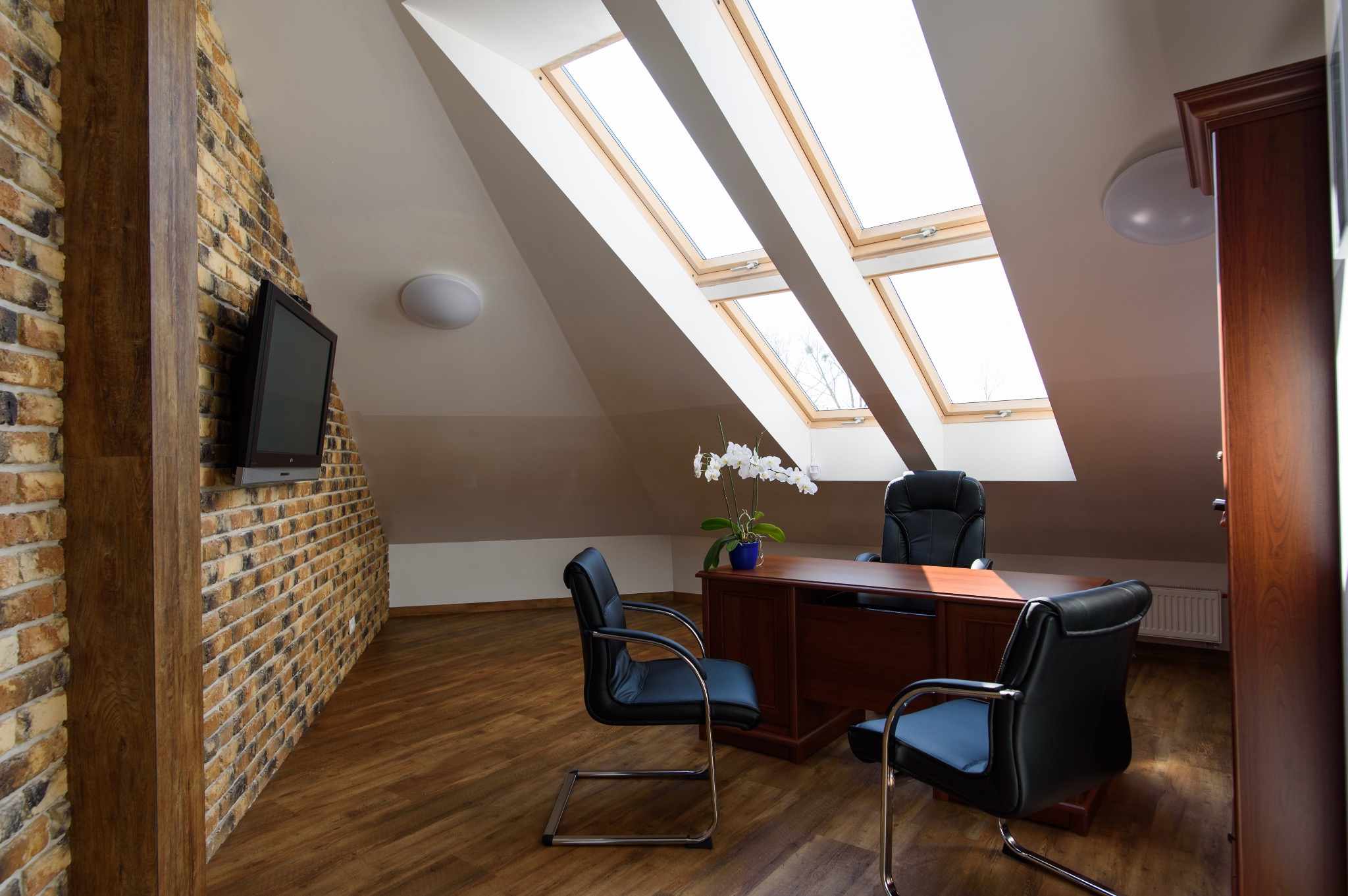 Jasne biuro z oknami dachowymi, biurkiem, krzesłami i telewizorem na ceglanej ścianie. Wazon z białymi kwiatami na biurku. Przytulne wnętrze.