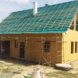 Dom w stanie surowym otwartym, ściany obłożone płytami drewnopochodnymi, dach pokryty zieloną membraną, widoczne rusztowania i tablica informacyjna budowy.