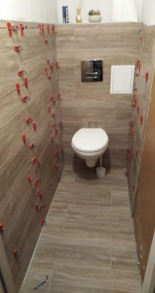 Układanie płytek w małej łazience z użyciem klipsów poziomujących, widoczna toaleta i zabudowa geberit, na półce dekoracje.