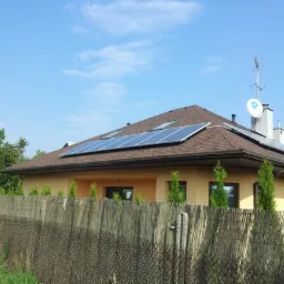 Dom jednorodzinny z brązowym dachem, na którym zamontowano panele słoneczne; widoczna antena satelitarna i ogrodzenie z siatki.