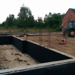 Wykop pod fundament z zabezpieczonymi ścianami, wystającymi prętami zbrojeniowymi oraz ubijarką i koparką w tle.