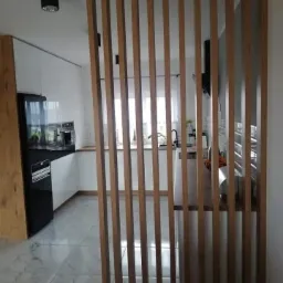 Drewniana ażurowa ścianka działowa oddzielająca kuchnię z białymi frontami i czarnymi akcentami od reszty pomieszczenia, widok z niskiej perspektywy.