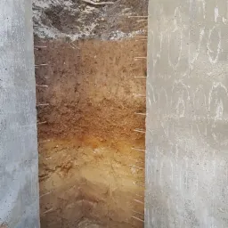 Wykop w ziemi z widocznymi warstwami gleby, zabezpieczony betonowymi ścianami z wystającymi prętami zbrojeniowymi.