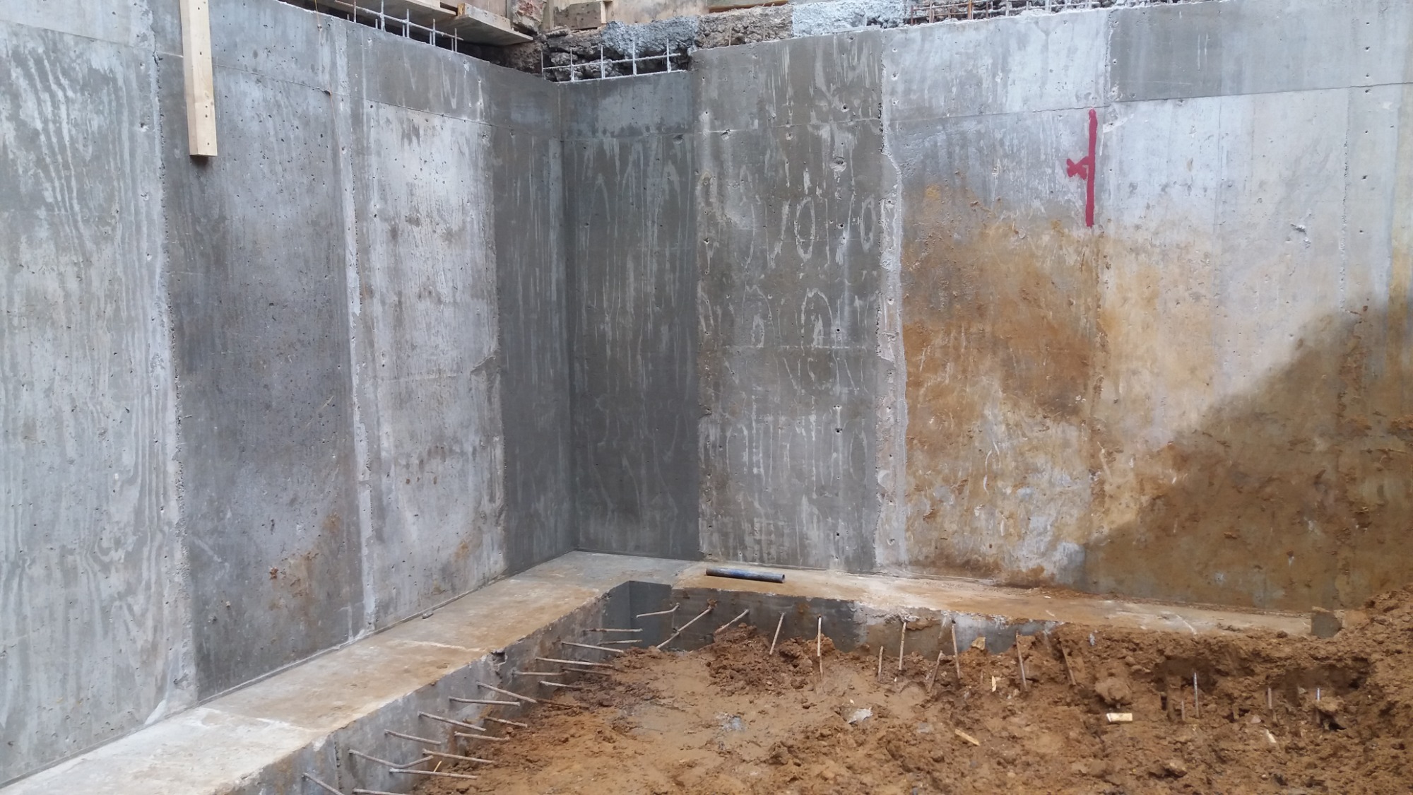 Surowe ściany fundamentowe z betonu z widocznymi śladami szalunku, częściowo zasypane ziemią, z wystającymi prętami zbrojeniowymi przygotowane do dalszych prac.