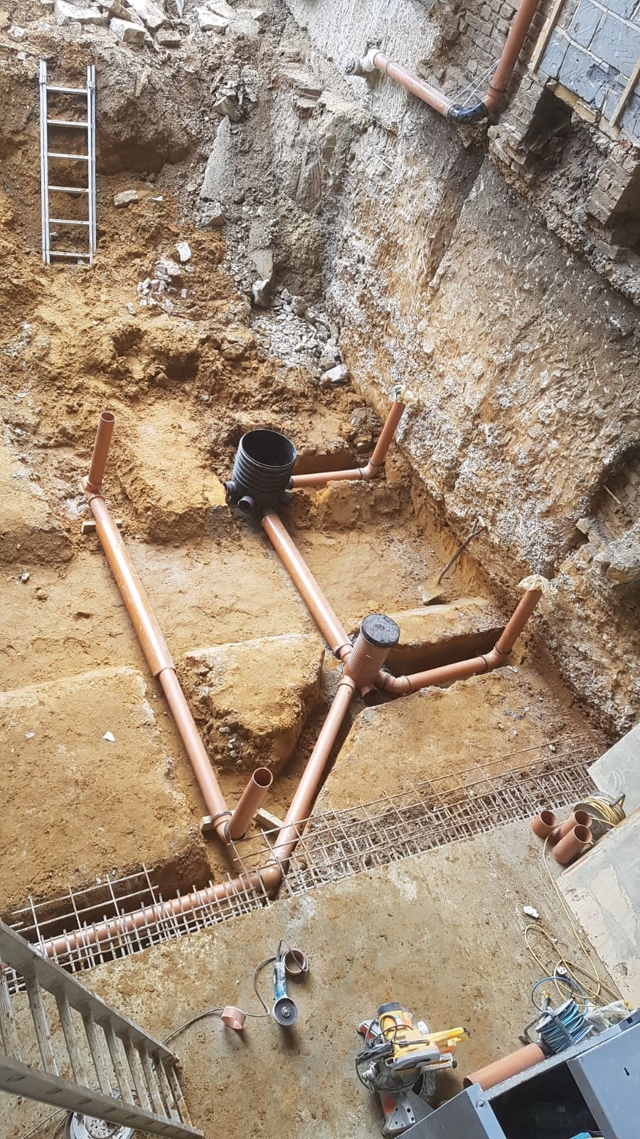 Wykop z widoczną instalacją rur kanalizacyjnych z tworzywa sztucznego, studzienki rewizyjne, zbrojenie i drabina oparta o ścianę wykopu, w tle ściana budynku z cegły.