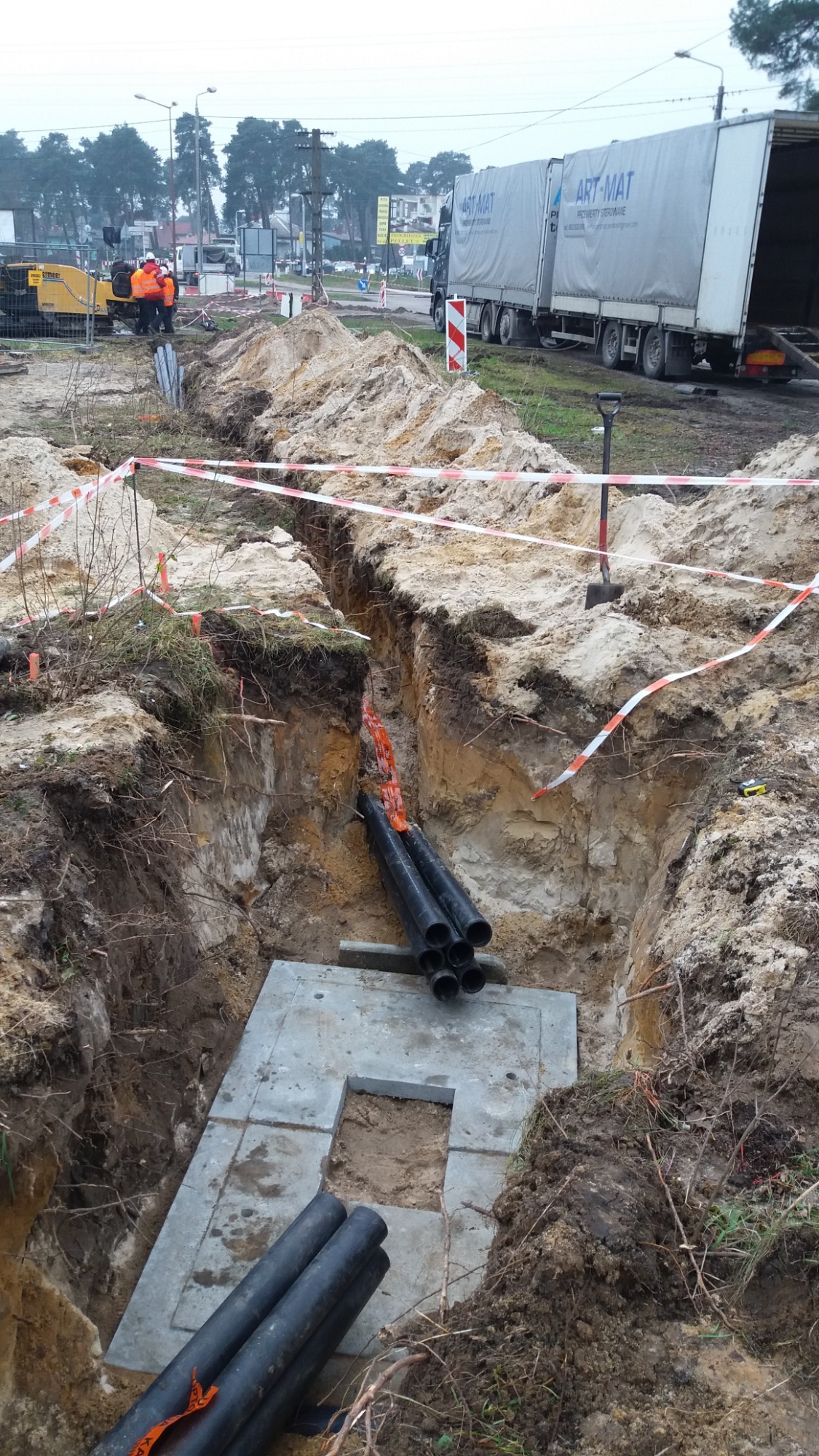 Wykop z betonowym elementem i rurami kanalizacyjnymi w ziemi, zabezpieczony taśmą ostrzegawczą, w tle widoczny sprzęt budowlany i ciężarówka.
