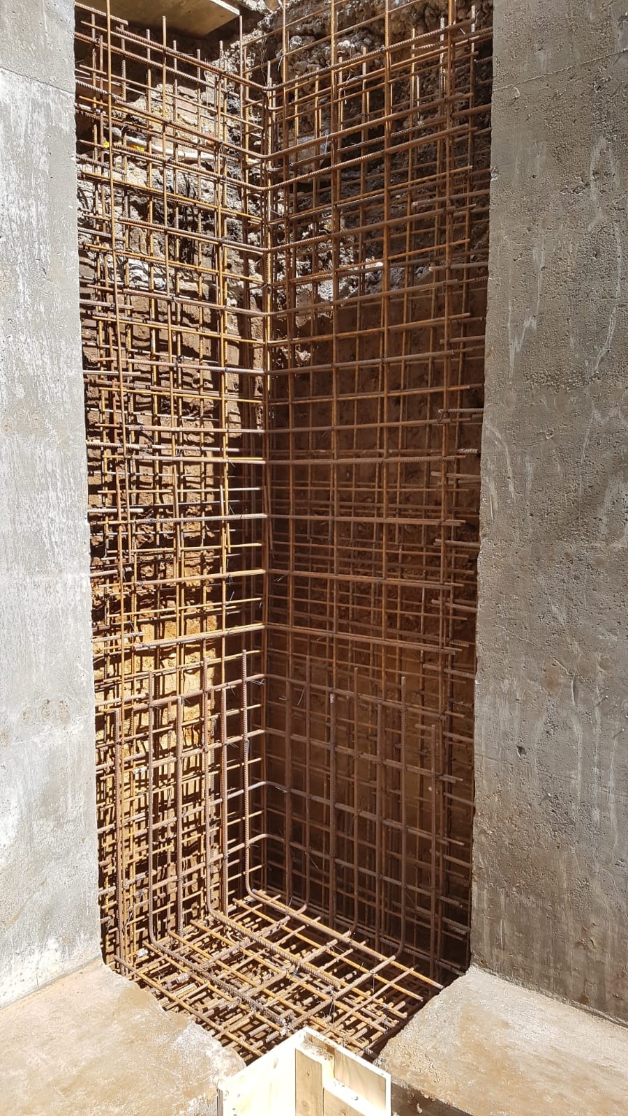 Szkielet z prętów zbrojeniowych w wykopie, przygotowany do zalania betonem. Widoczne szalunki betonowe po bokach i fragment drewnianych elementów szalunkowych na dole.