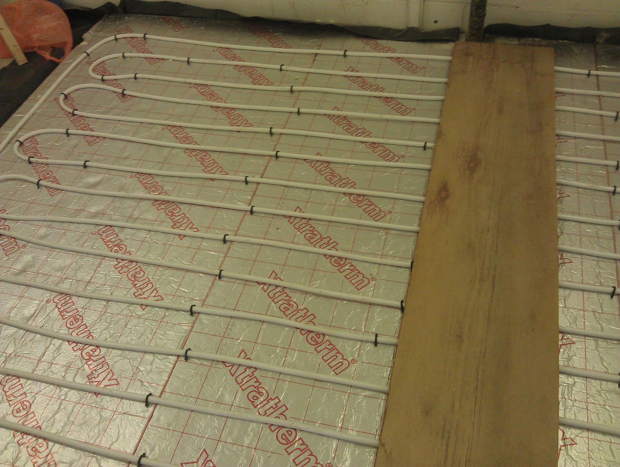 Białe rurki ogrzewania podłogowego ułożone na srebrnej folii z czerwonym nadrukiem, częściowo przykryte drewnianą płytą.