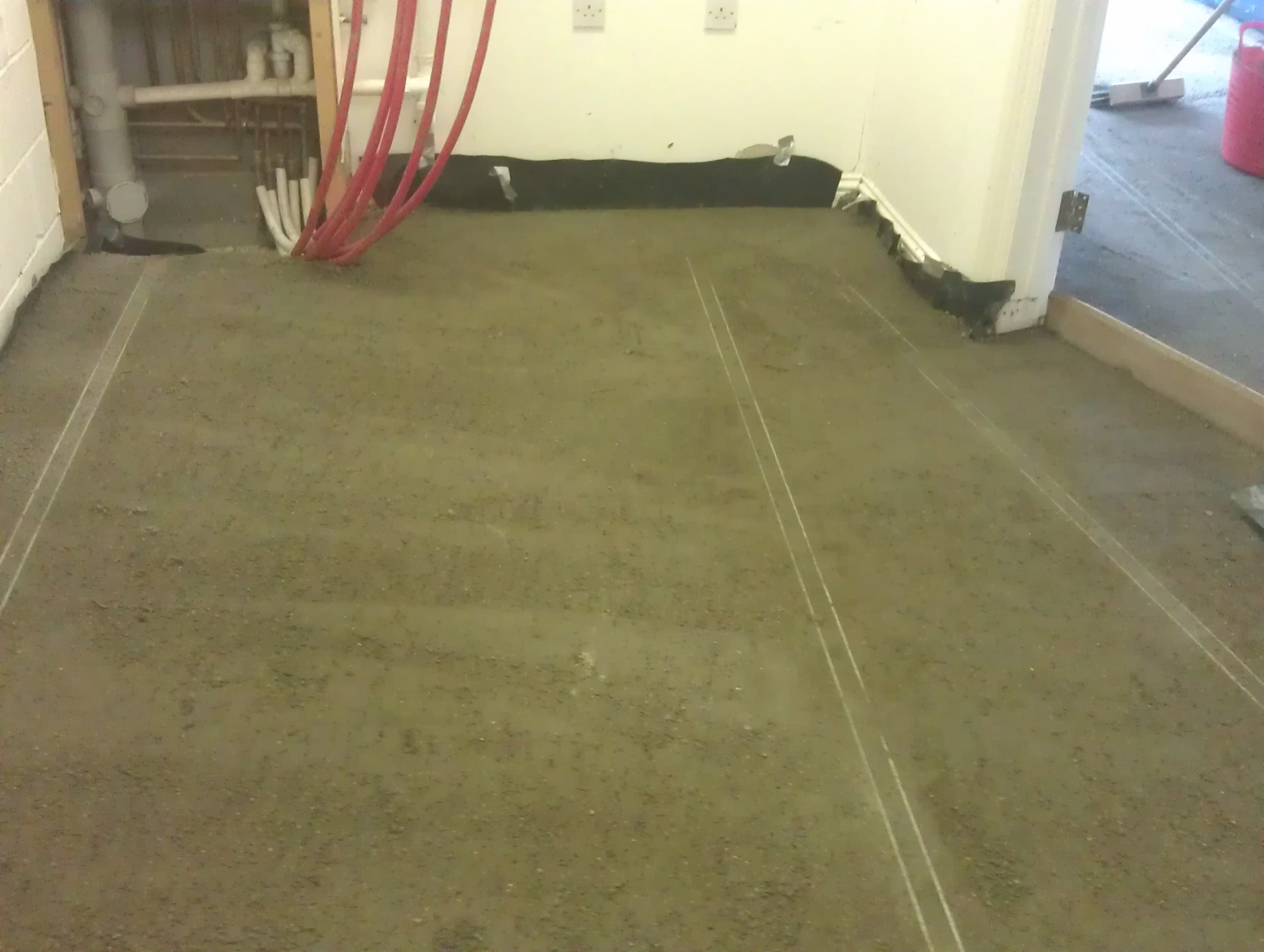 Wyrównywanie podłoża w pomieszczeniu z instalacją wodną; widoczne czerwone rury, listwy przypodłogowe i cienkie białe linie wyznaczające obszar roboczy.