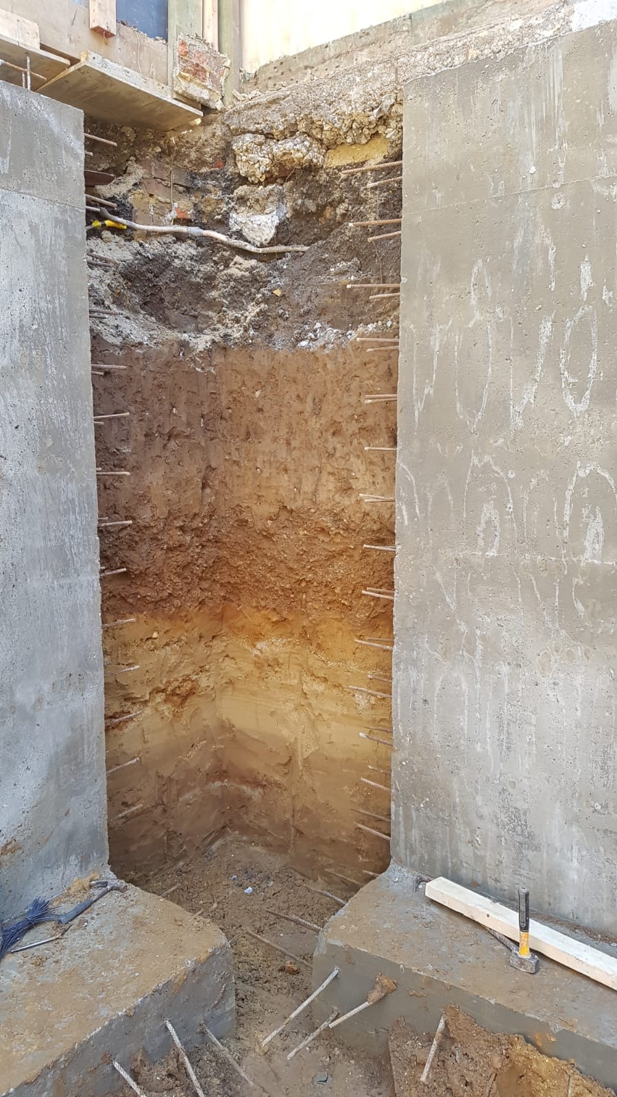 Wykop w ziemi z widocznymi warstwami gleby, zabezpieczony betonowymi ścianami z wystającymi prętami zbrojeniowymi.