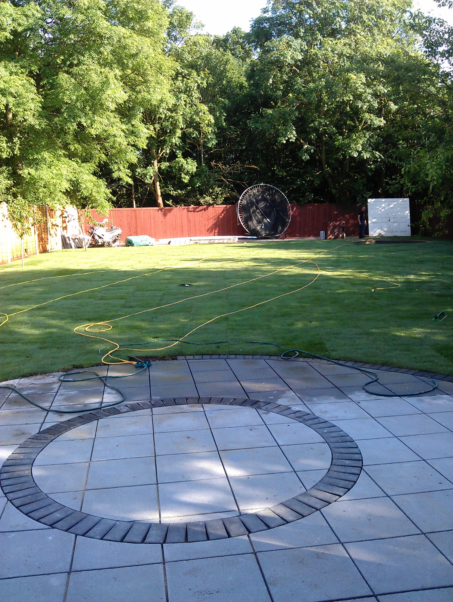 Wykończony taras z kostki brukowej w ogrodzie z widocznym trawnikiem, ogrodzeniem i trampoliną w tle.