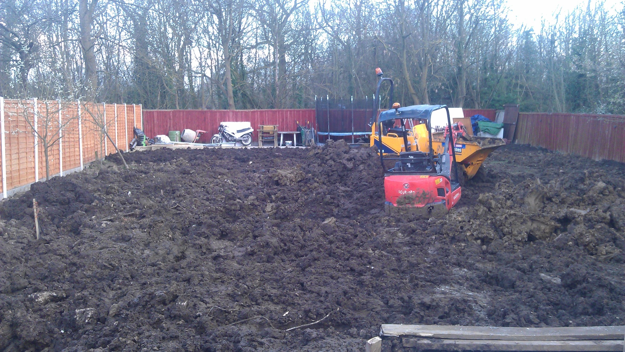 Czerwony minikoparka Kubota i żółta wywrotka na grząskim, błotnistym terenie w ogrodzie, obok stoją zaparkowany skuter i trampolina