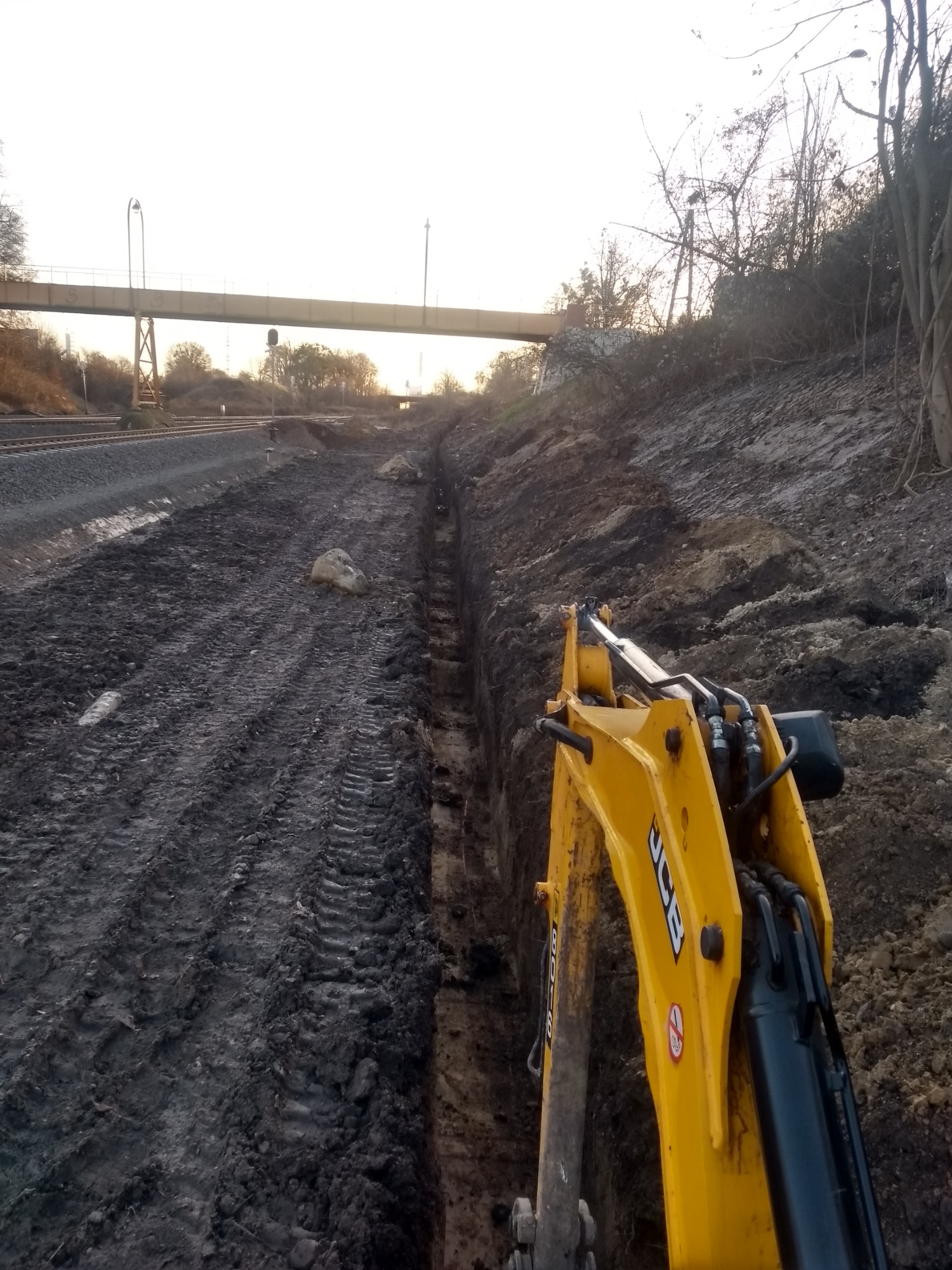 Żółta koparka JCB wykopuje wąski rów w ziemi, obok torów kolejowych i wiaduktu w tle.
