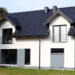 Nowoczesny, dwukondygnacyjny dom z białymi ścianami, czarną dachówką i szarymi akcentami, z widocznymi panelami słonecznymi na dachu sąsiedniego budynku.